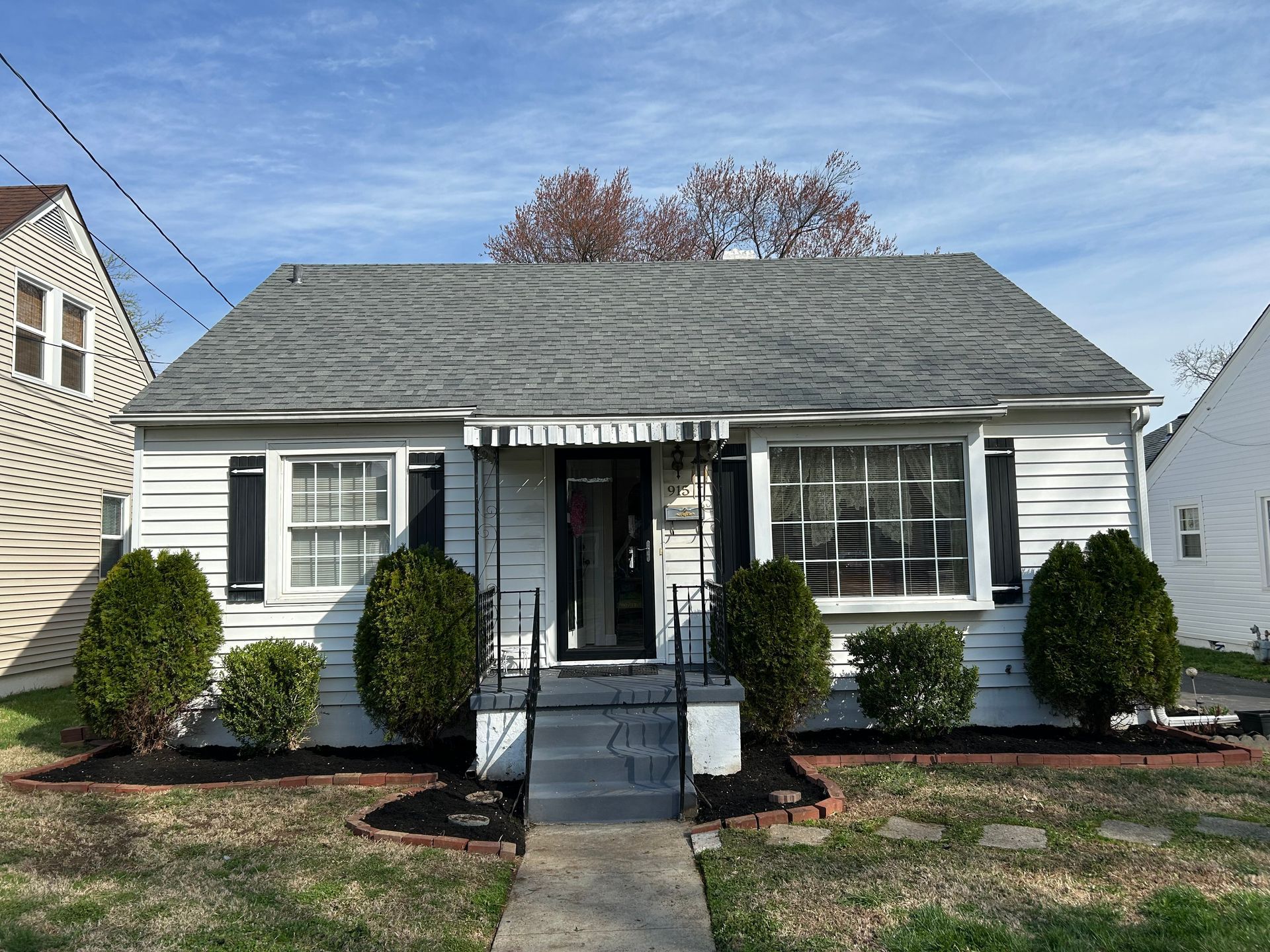 Small white single-story house with gray roof, black shutters, and a front porch with steps and shrubs.