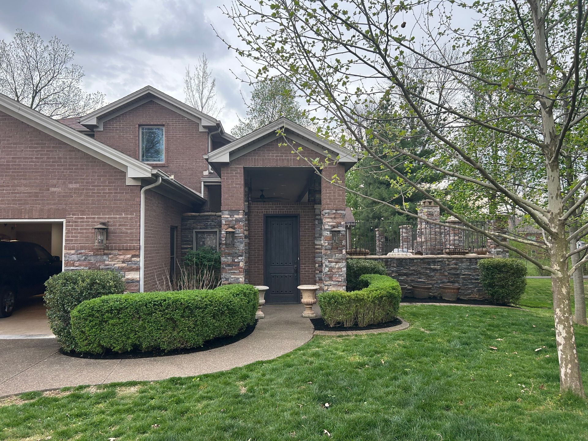 Brick suburban house with a front porch, trimmed hedges, and a green lawn under cloudy skies