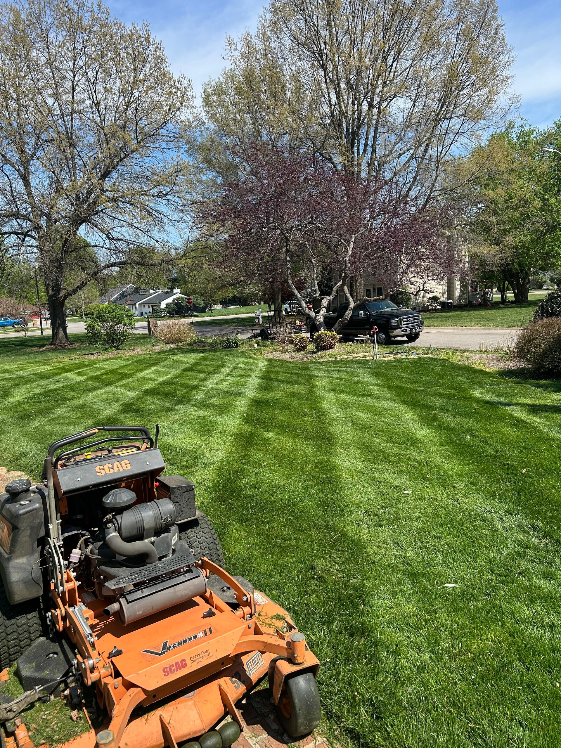 Orange riding lawn mower on a green striped lawn in a sunny park with trees and parked cars.