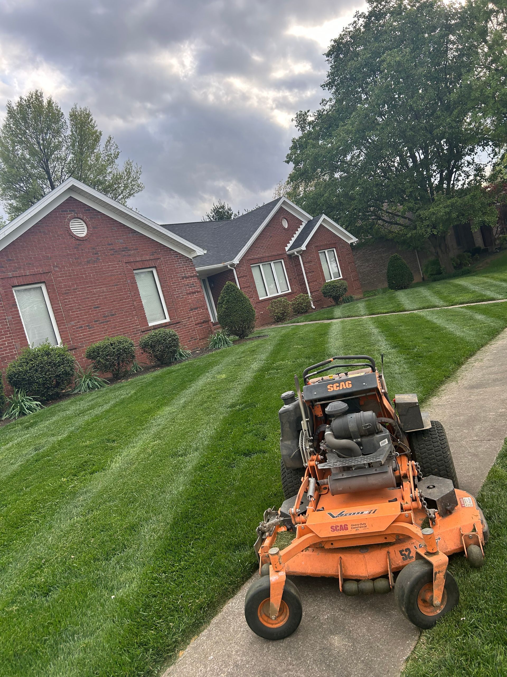 Orange riding lawn mower on a steep grassy lawn beside red brick houses under cloudy sky