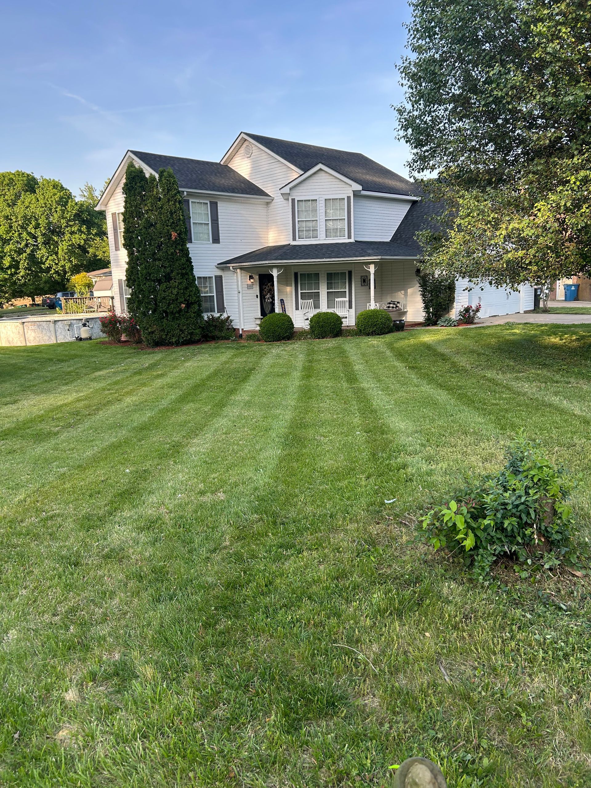 White two-story house with a front porch, large green lawn, and trees under a blue sky