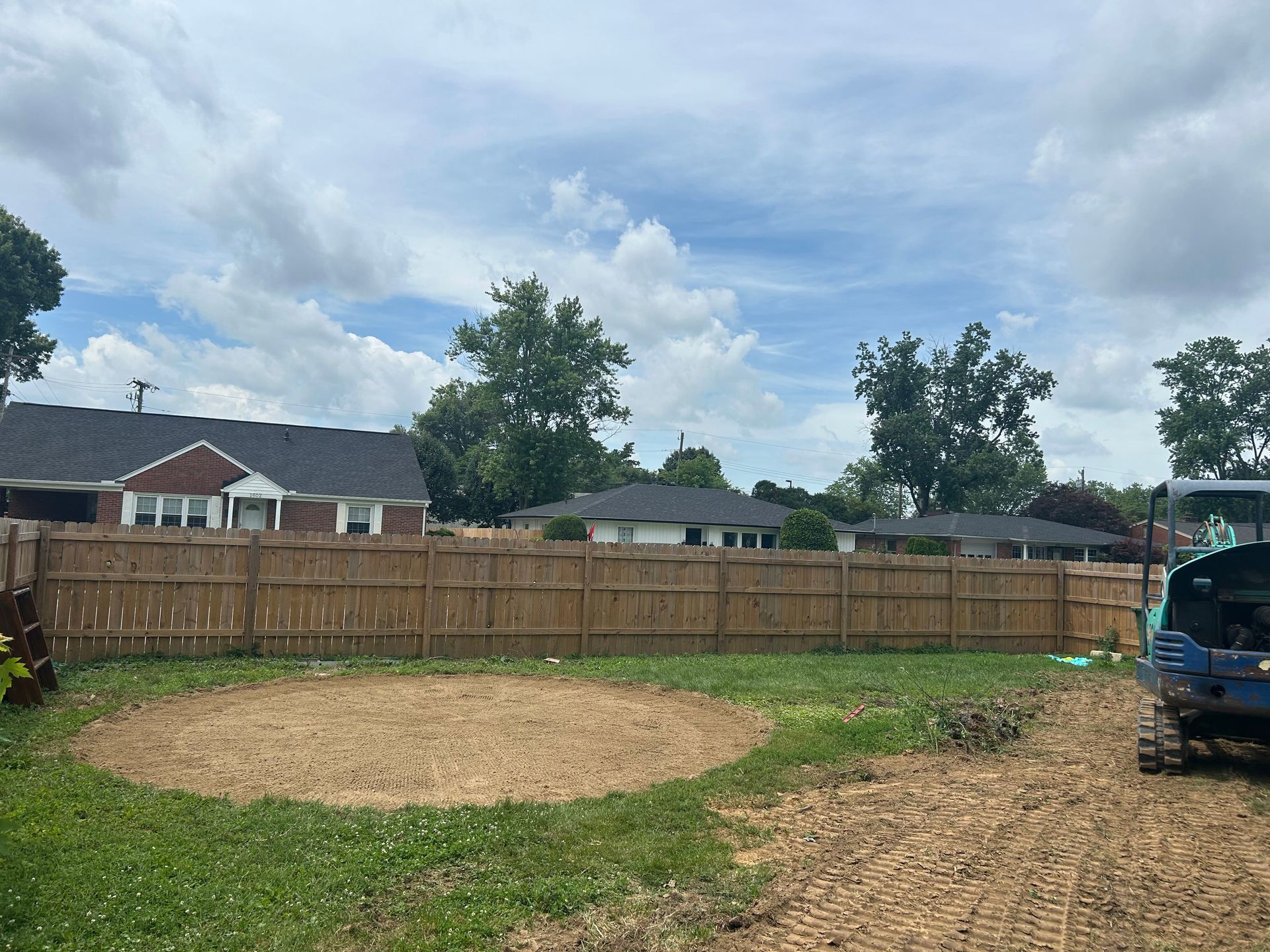 Backyard with fenced yard, bare dirt patch, gravel path, trees, and parked car under cloudy sky