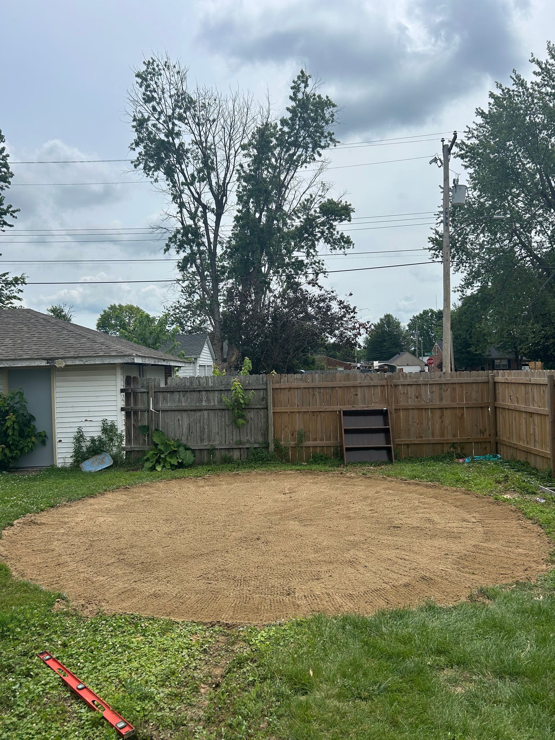 Backyard with a large gravel patch, wooden fence, trees, and a small red tool in the grass.