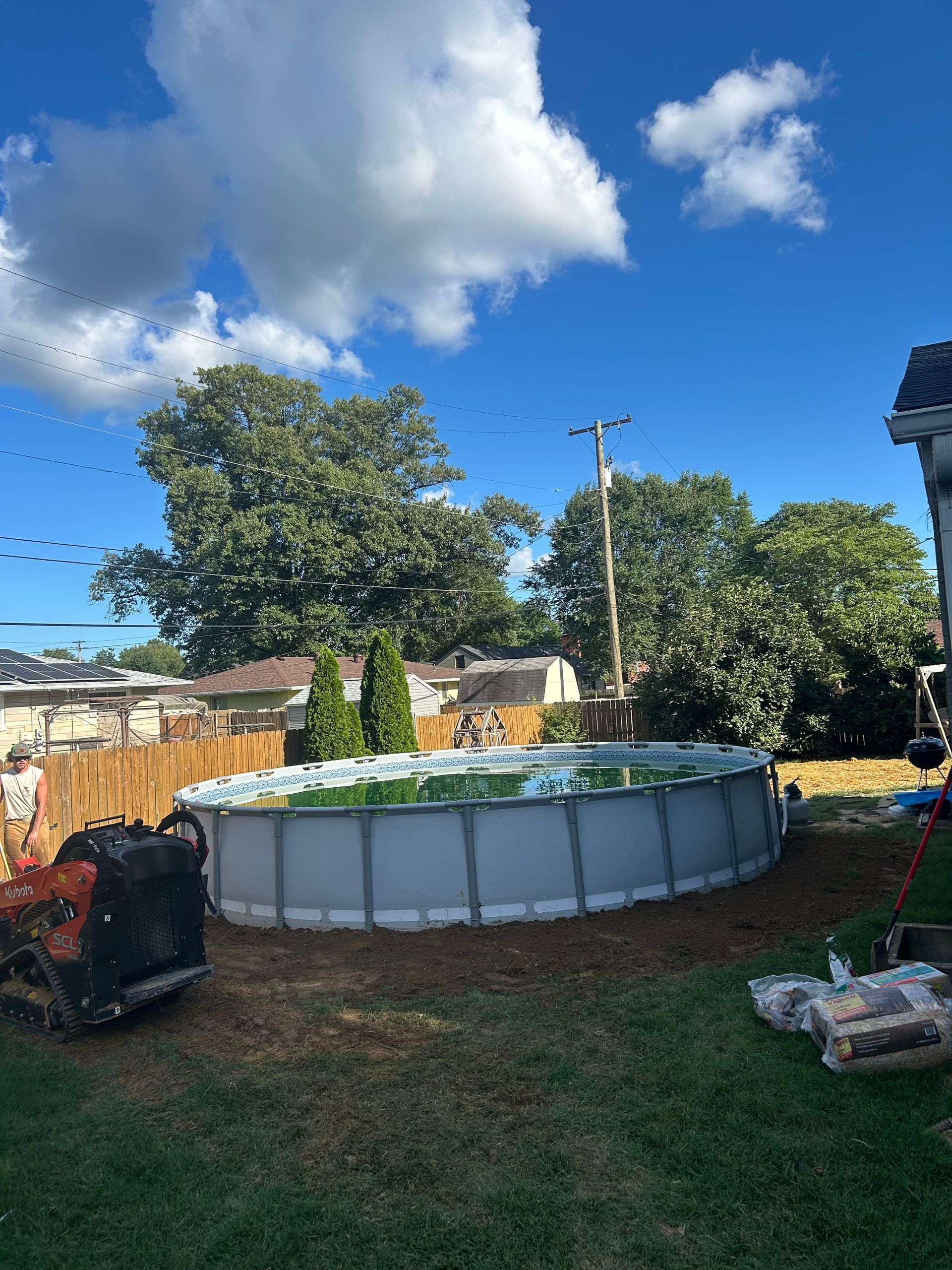 Backyard with an above-ground pool, grassy lawn, trees, fence, and a bright blue sky with clouds