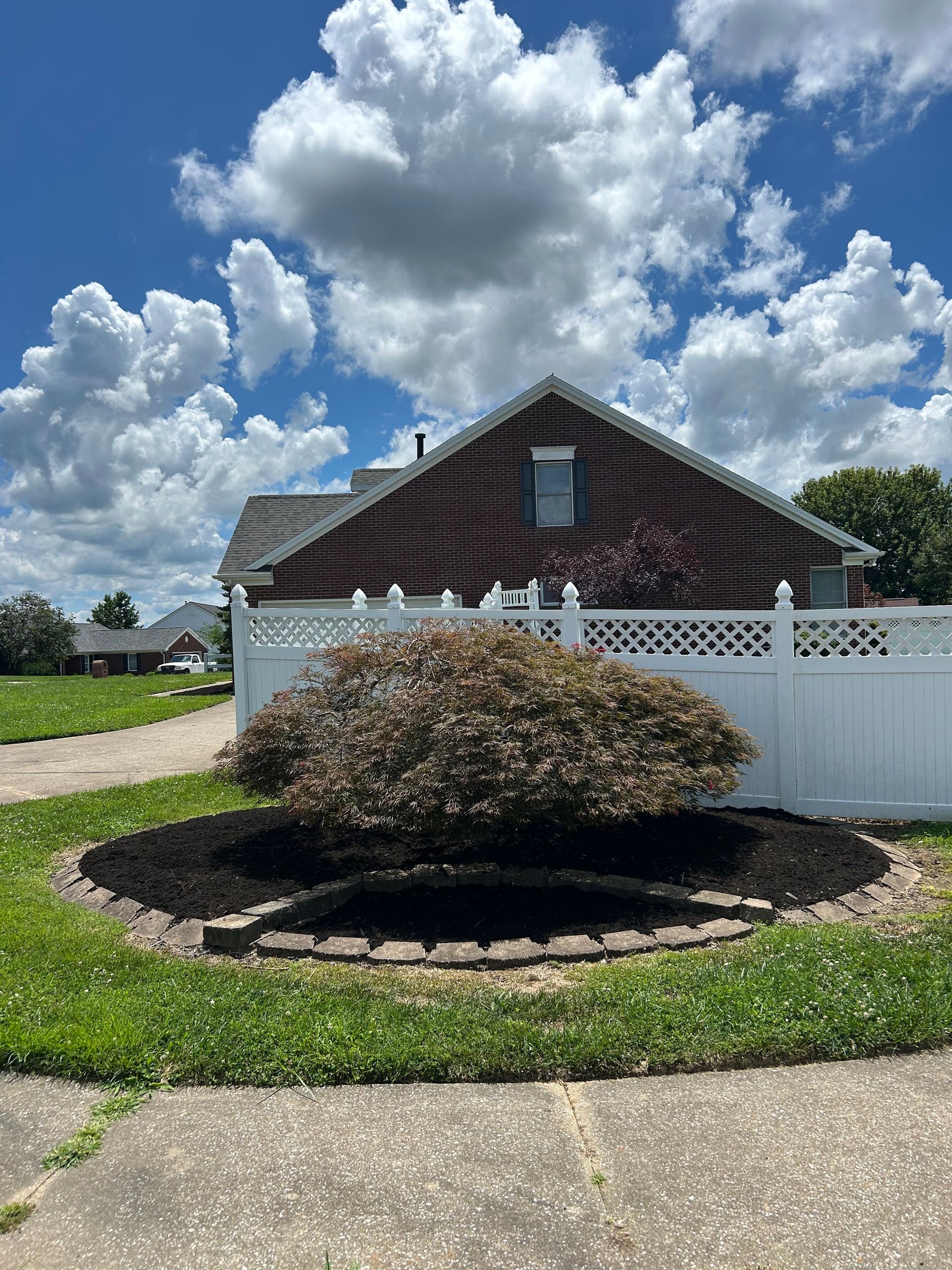 Suburban house with white fence, round shrub, and cloudy blue sky over a driveway and lawn
