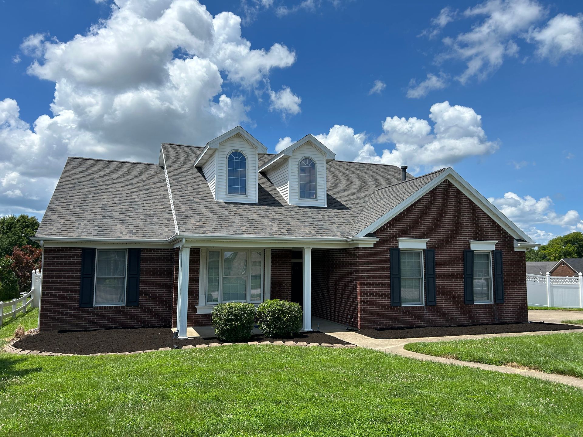 Suburban brick house with gray roof, dormer windows, and a green lawn under a blue sky.