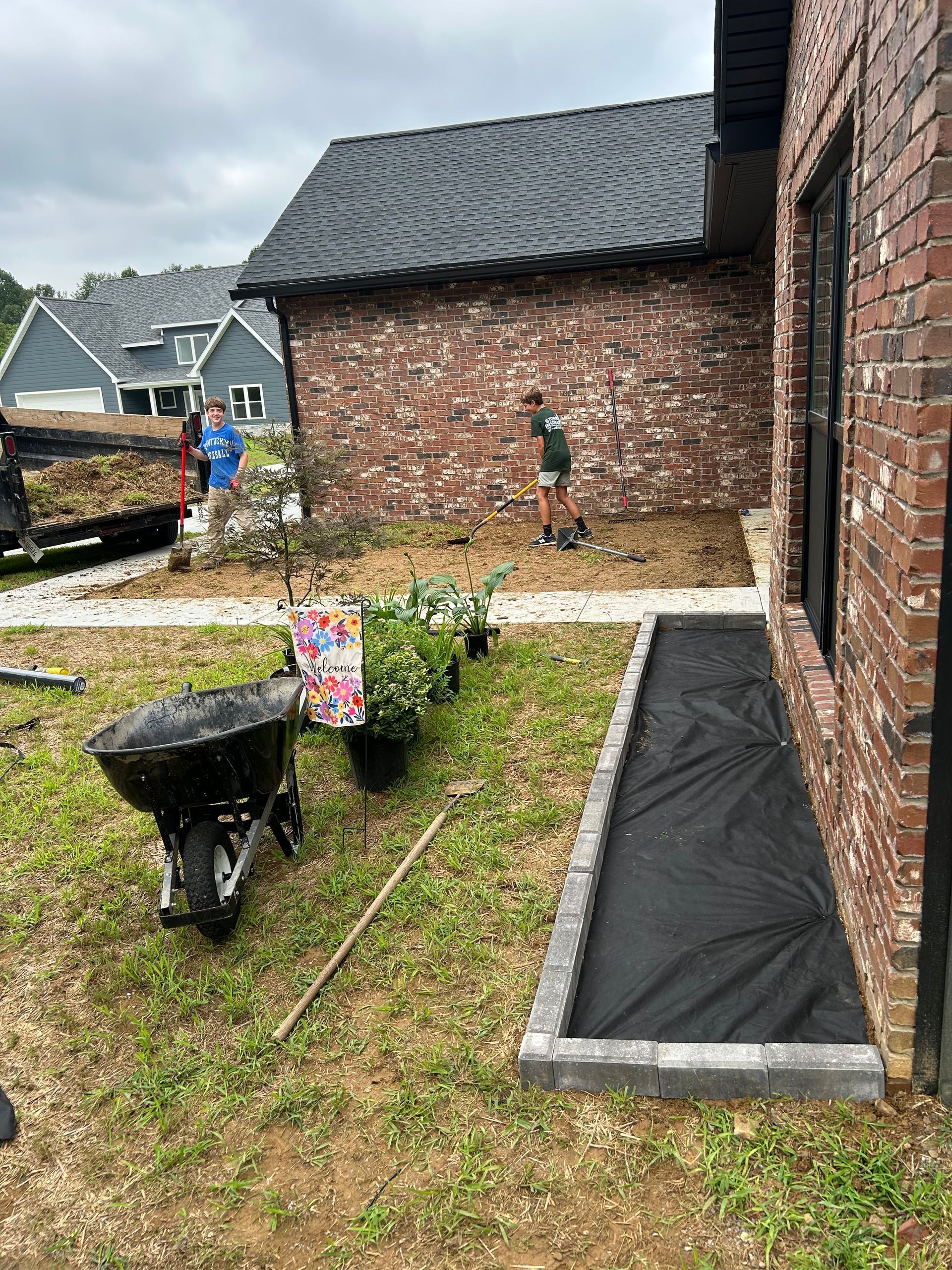 Two people working in a brick-walled garden with wheelbarrows, gravel path, and newly laid black edging