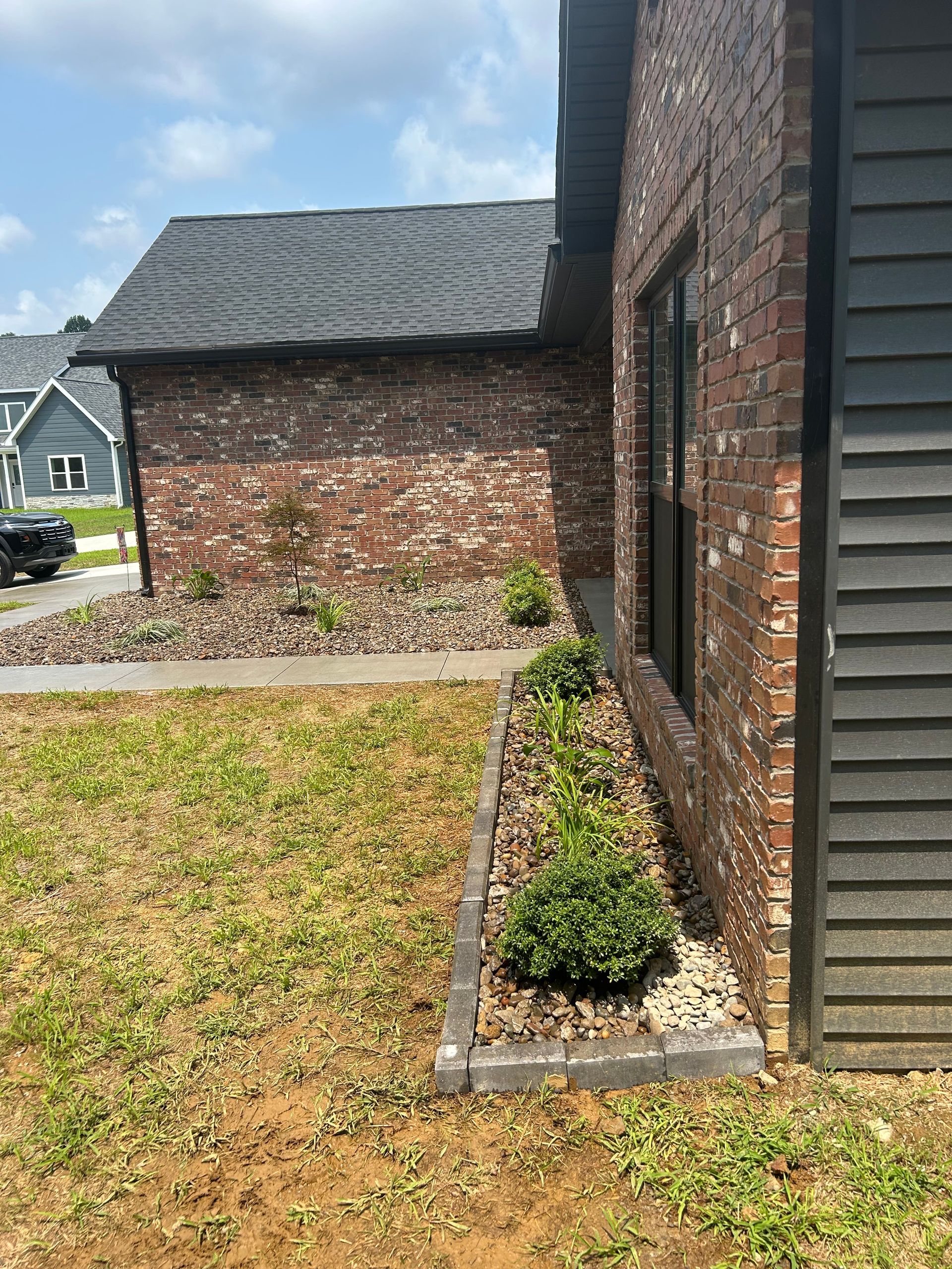 Brick house corner with a small shrub bed and grassy yard, neighboring brick garage in the background