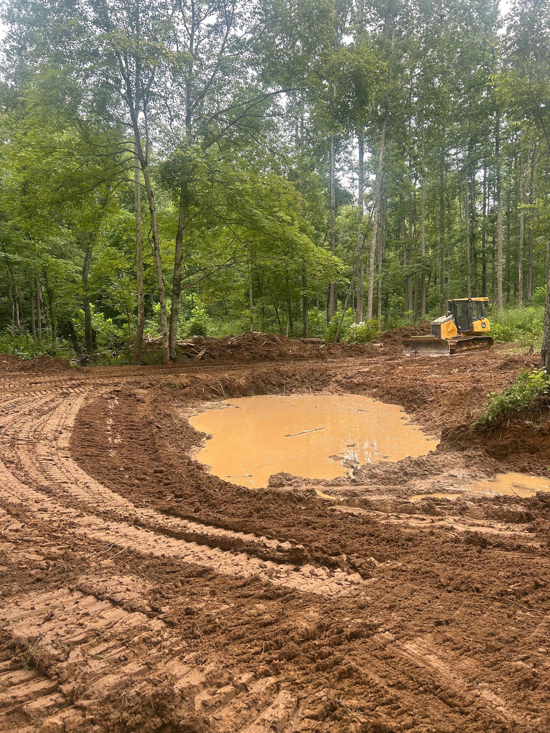 Mud-filled dirt track with a yellow bulldozer beside a muddy puddle in a wooded clearing