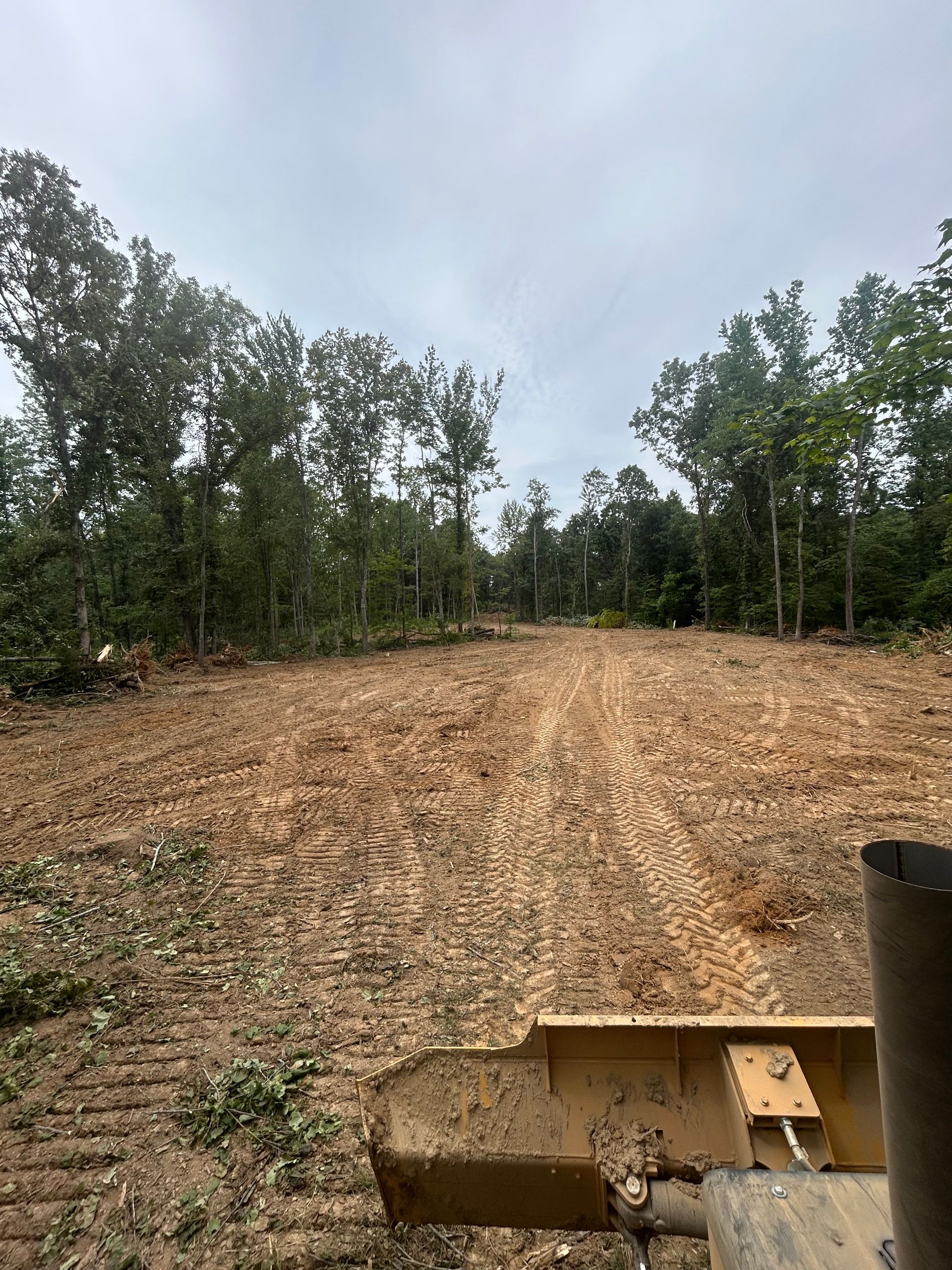 Cleared dirt path through trees in a wooded area, with part of a bulldozer visible in the foreground