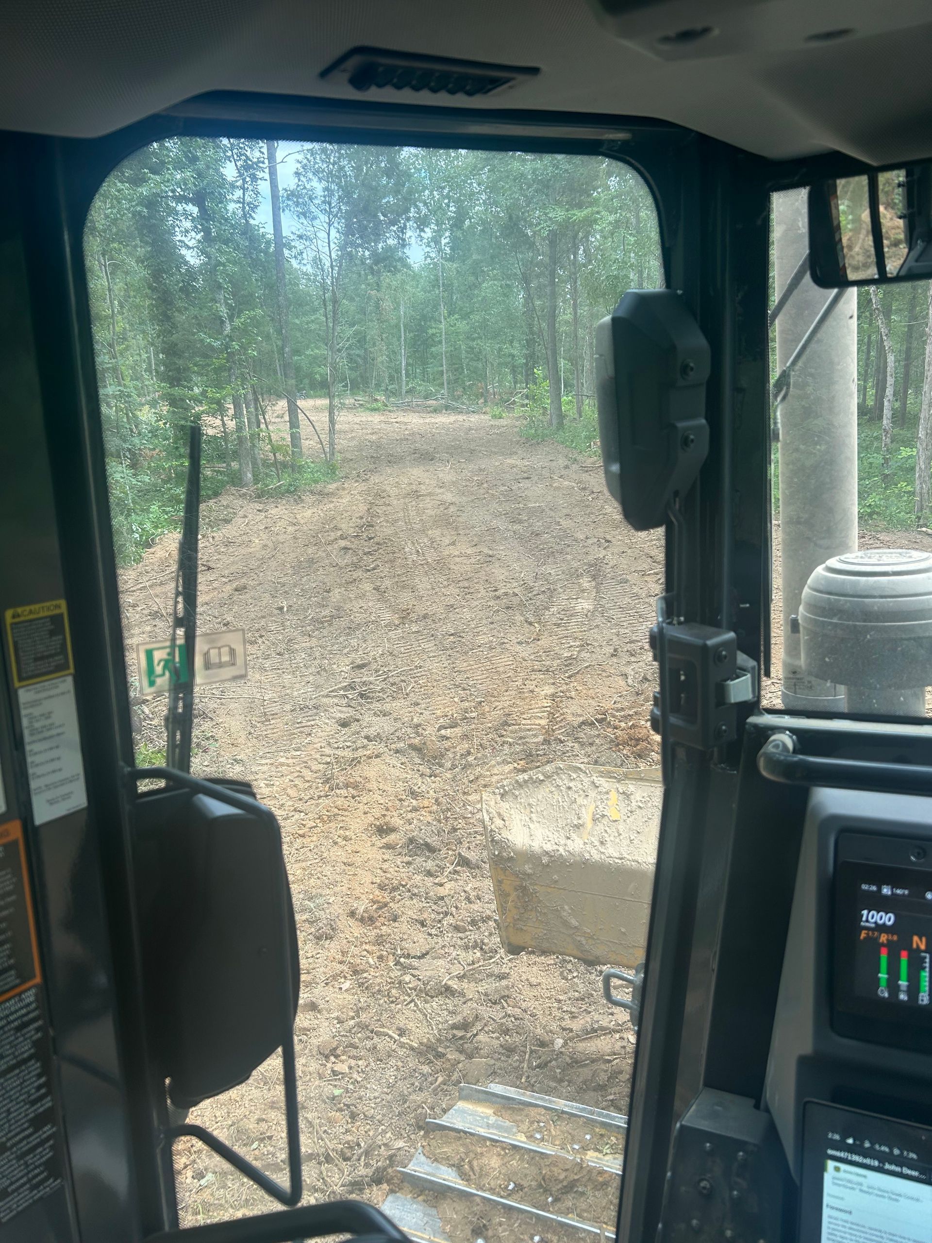 Inside a tractor cab looking out at a dirt path and trees in a wooded area.