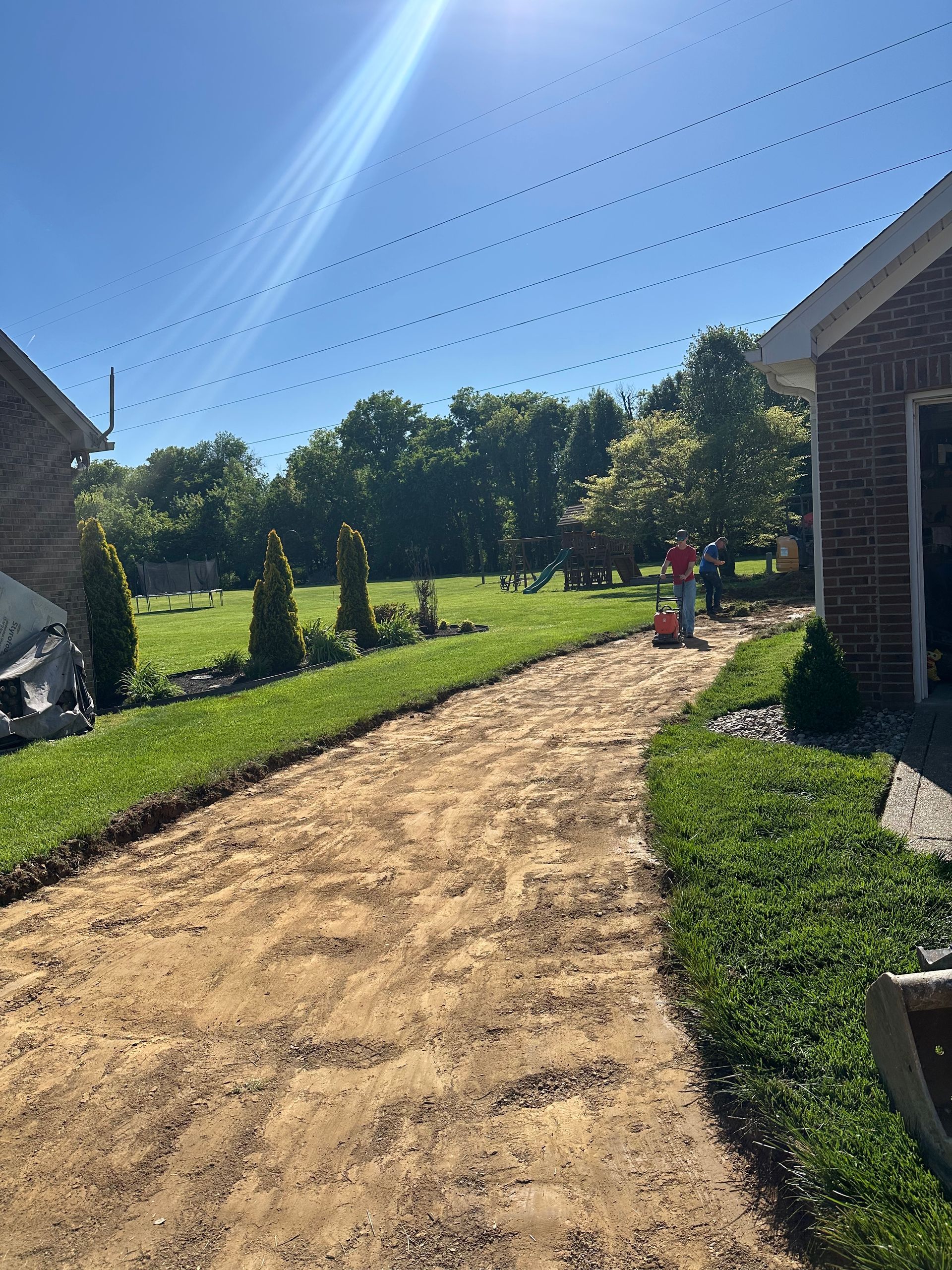 Sandy path through a sunny backyard with green lawn, trees, and a brick house under a clear blue sky