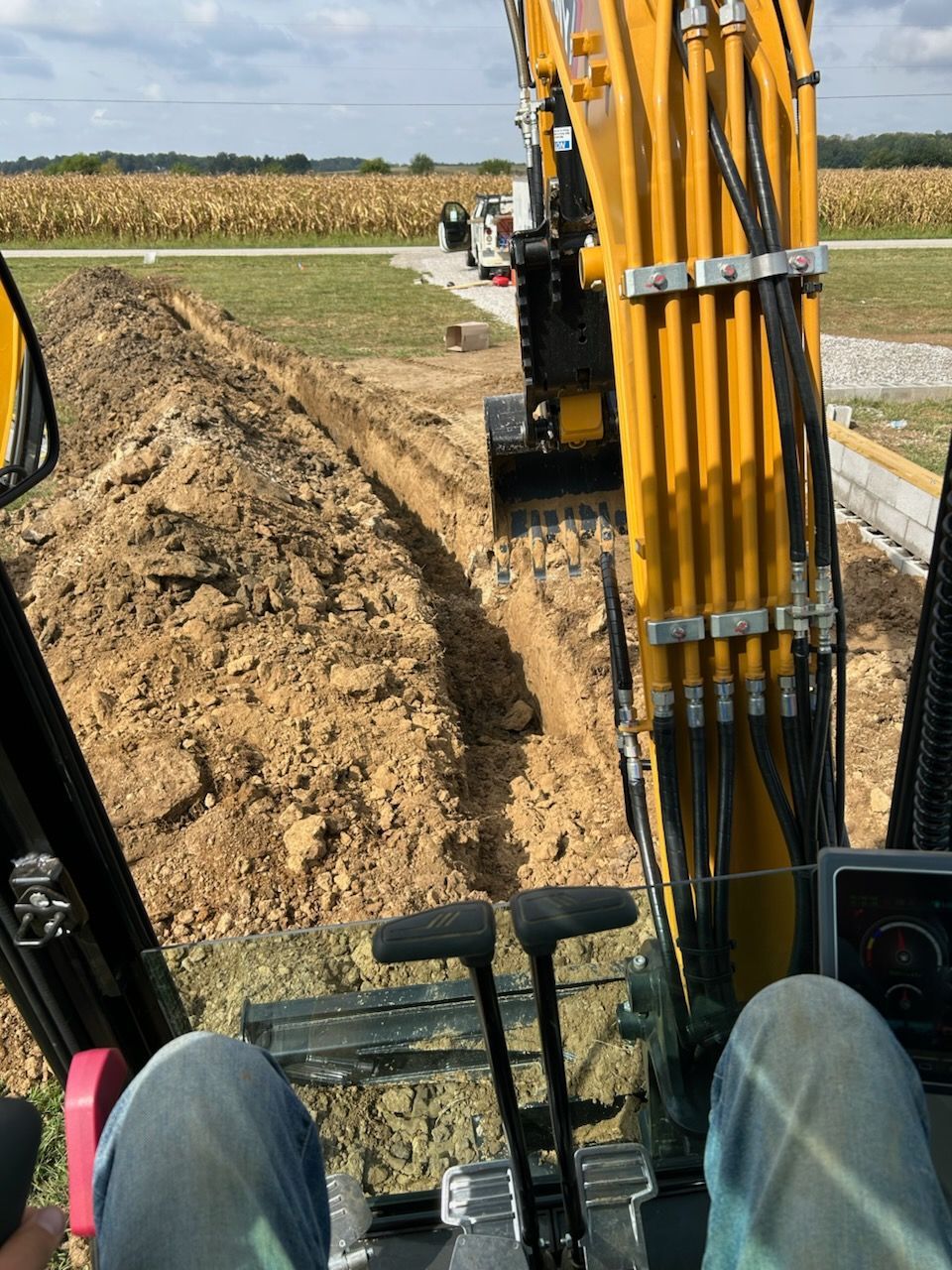 View from excavator cab of digging a dirt trench beside a field, with yellow boom in foreground