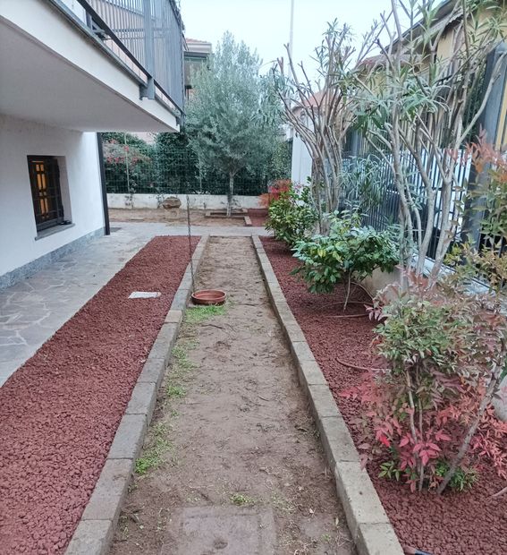 Stretto vialetto in giardino accanto a una casa, delimitato da pacciame rosso e arbusti, con terra nuda al centro.
