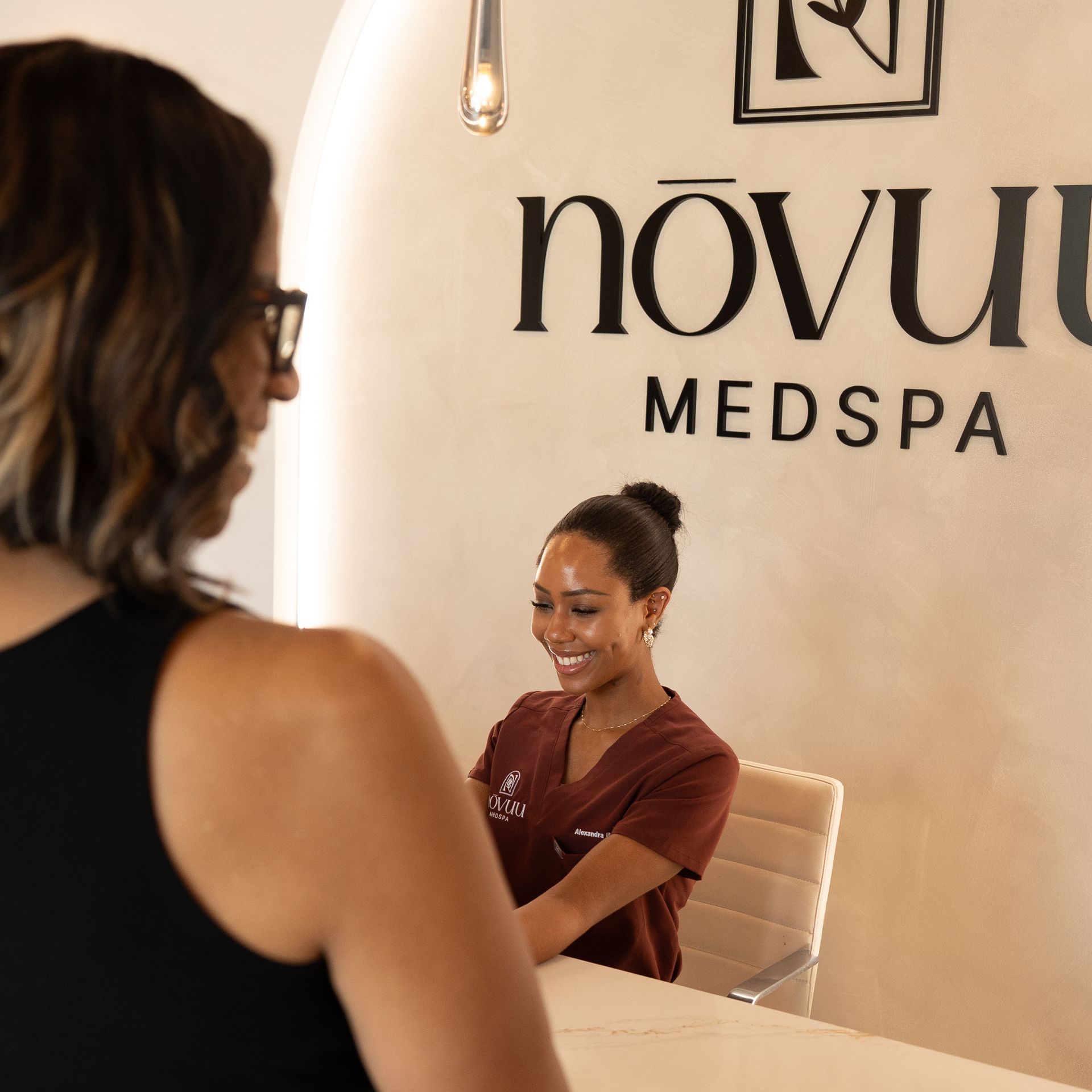 Two women talking in a bright med spa reception area with Novu Medspa sign on the wall