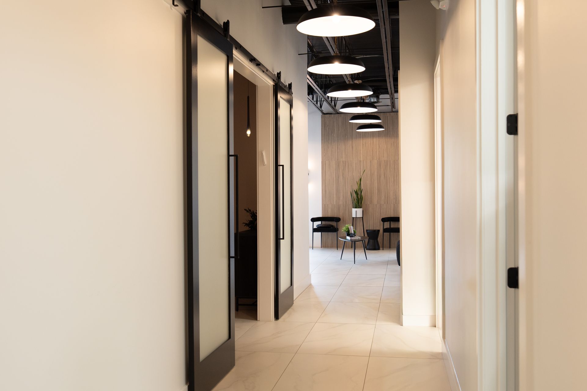 Bright office hallway with white walls, black doors, and pendant lights leading to a seating area.