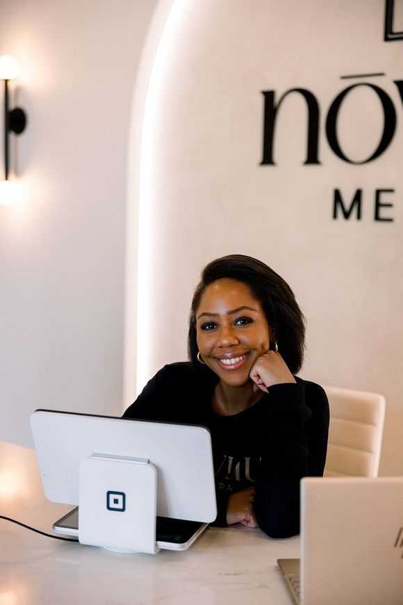 Smiling woman at desk with laptop in a bright office, seated in front of a wall logo.