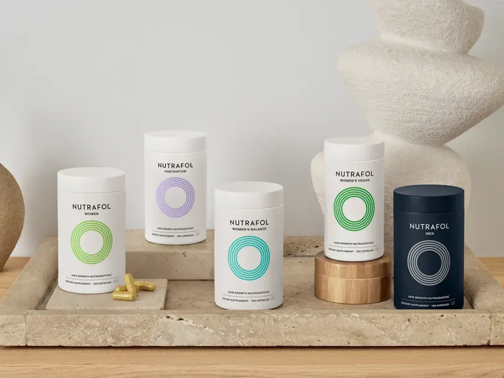 Five skincare jars in green, lavender, teal, white and navy arranged on beige stone with sculptural decor.
