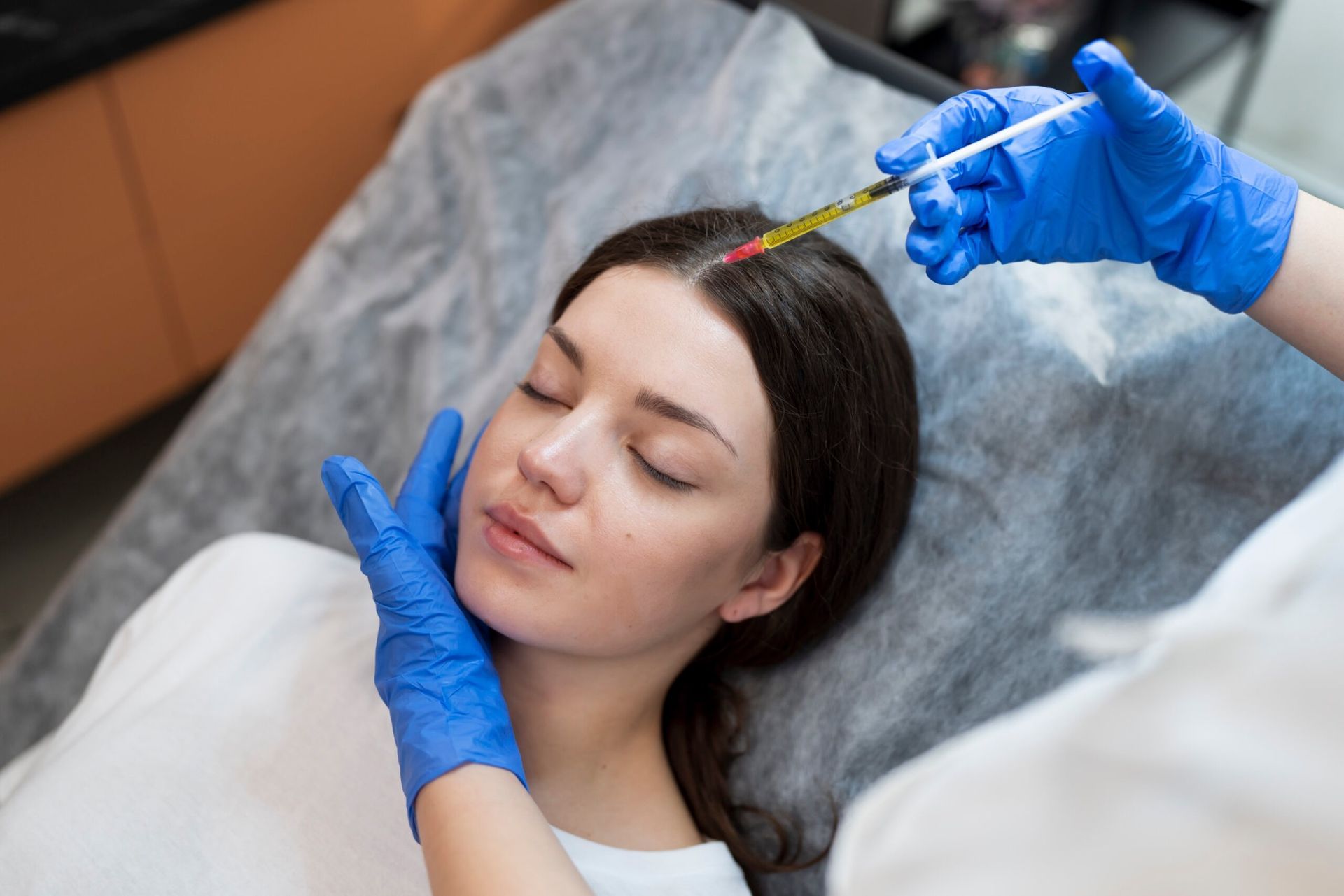 Cosmetic injection into forehead while patient reclines on treatment bed, gloved hands adjusting face