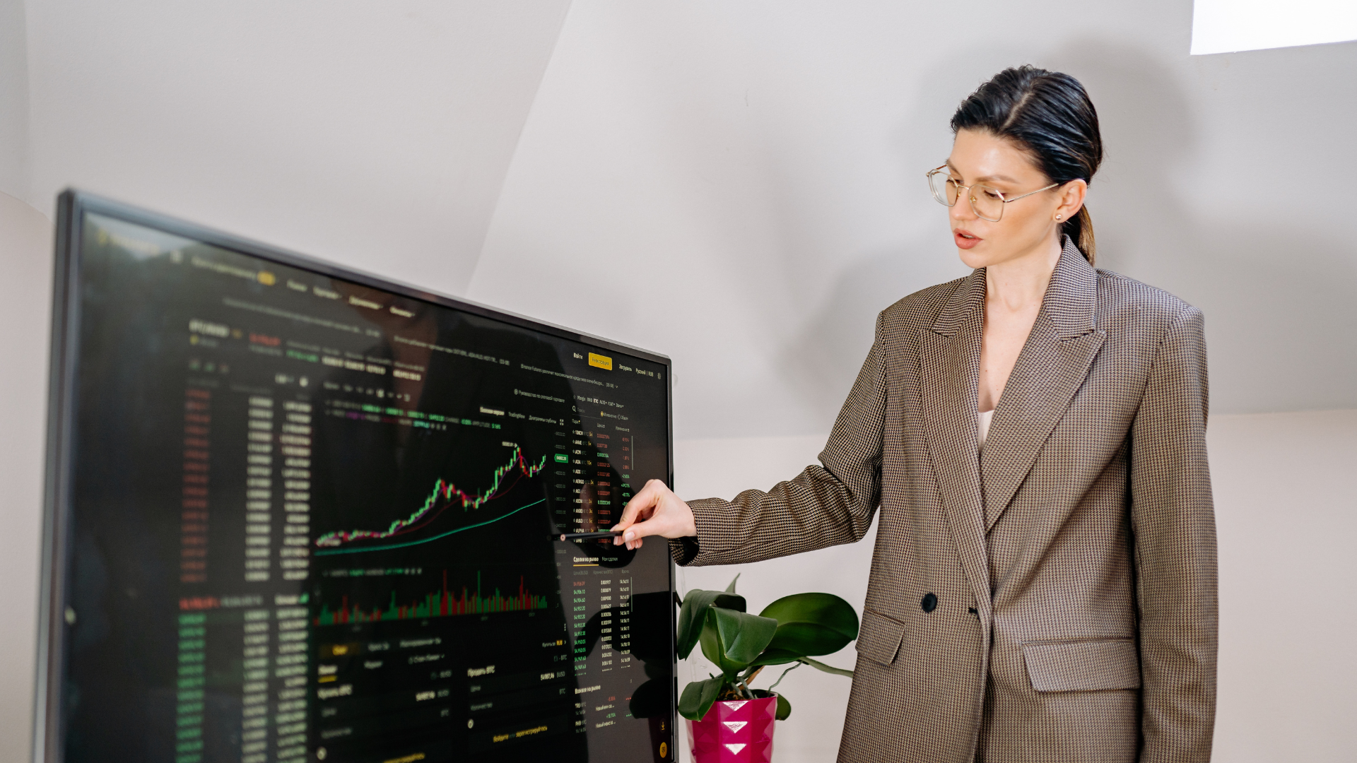 Person in a brown plaid suit pointing at a stock trading display with charts on a large monitor