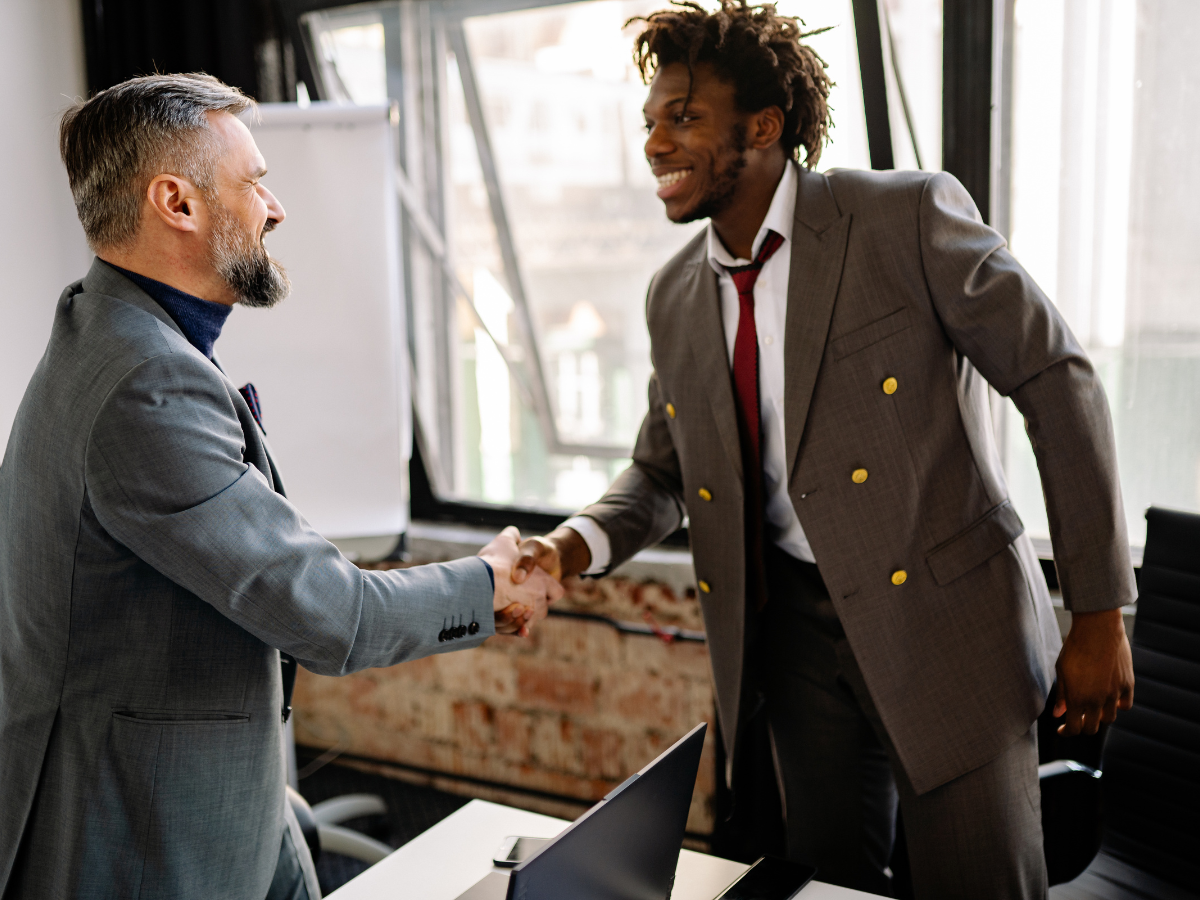 Two men in suits shaking hands in an office, smiling.