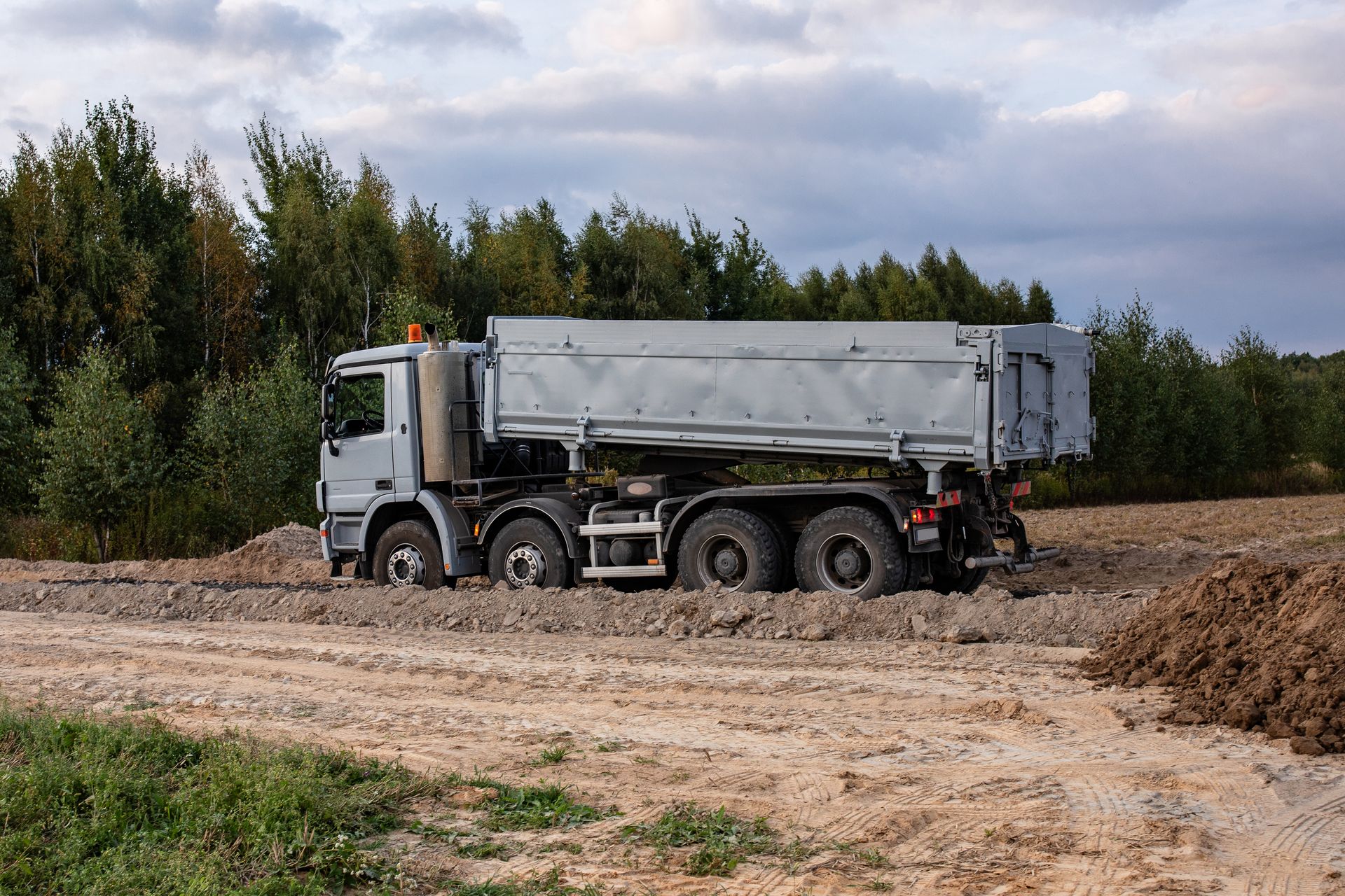 Gray dump truck stuck in muddy terrain near a tree line.