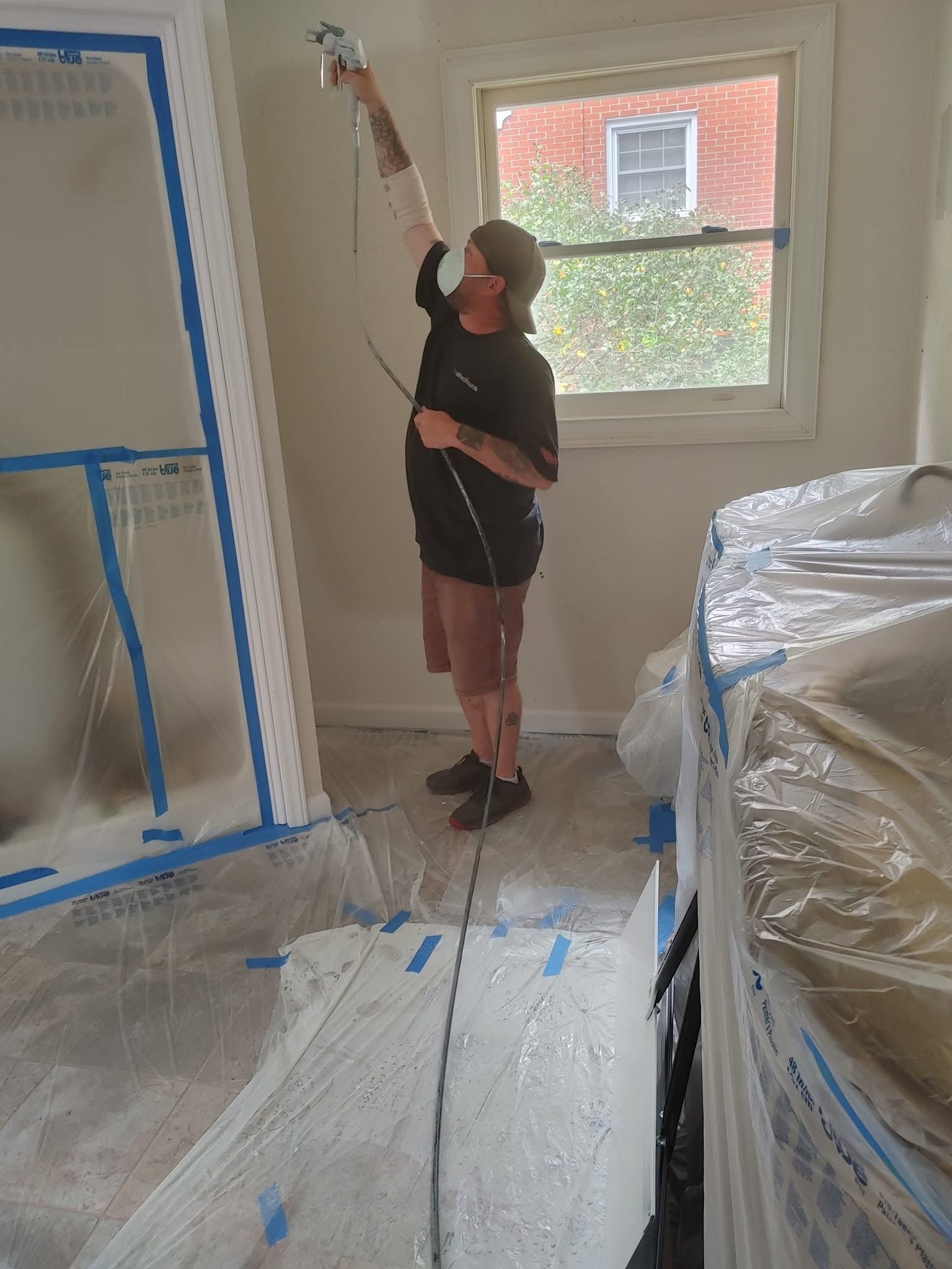 Person painting a wall with a roller in a room covered with protective plastic sheets.