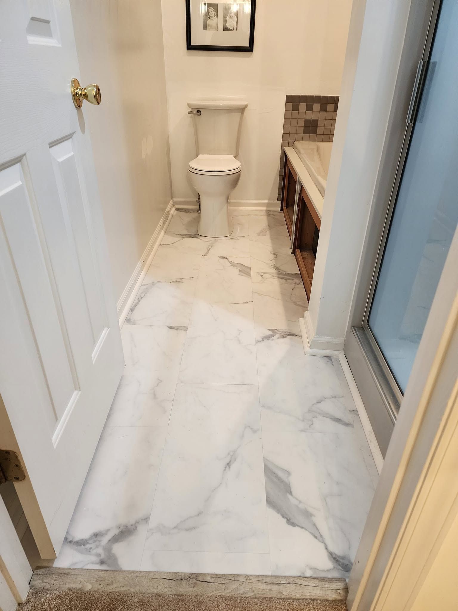 Narrow bathroom with white marble floor, toilet, vanity, and glass shower door.