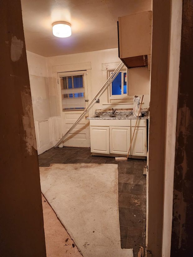 Damaged kitchen with torn flooring, exposed walls, and a white sink cabinet near a window.