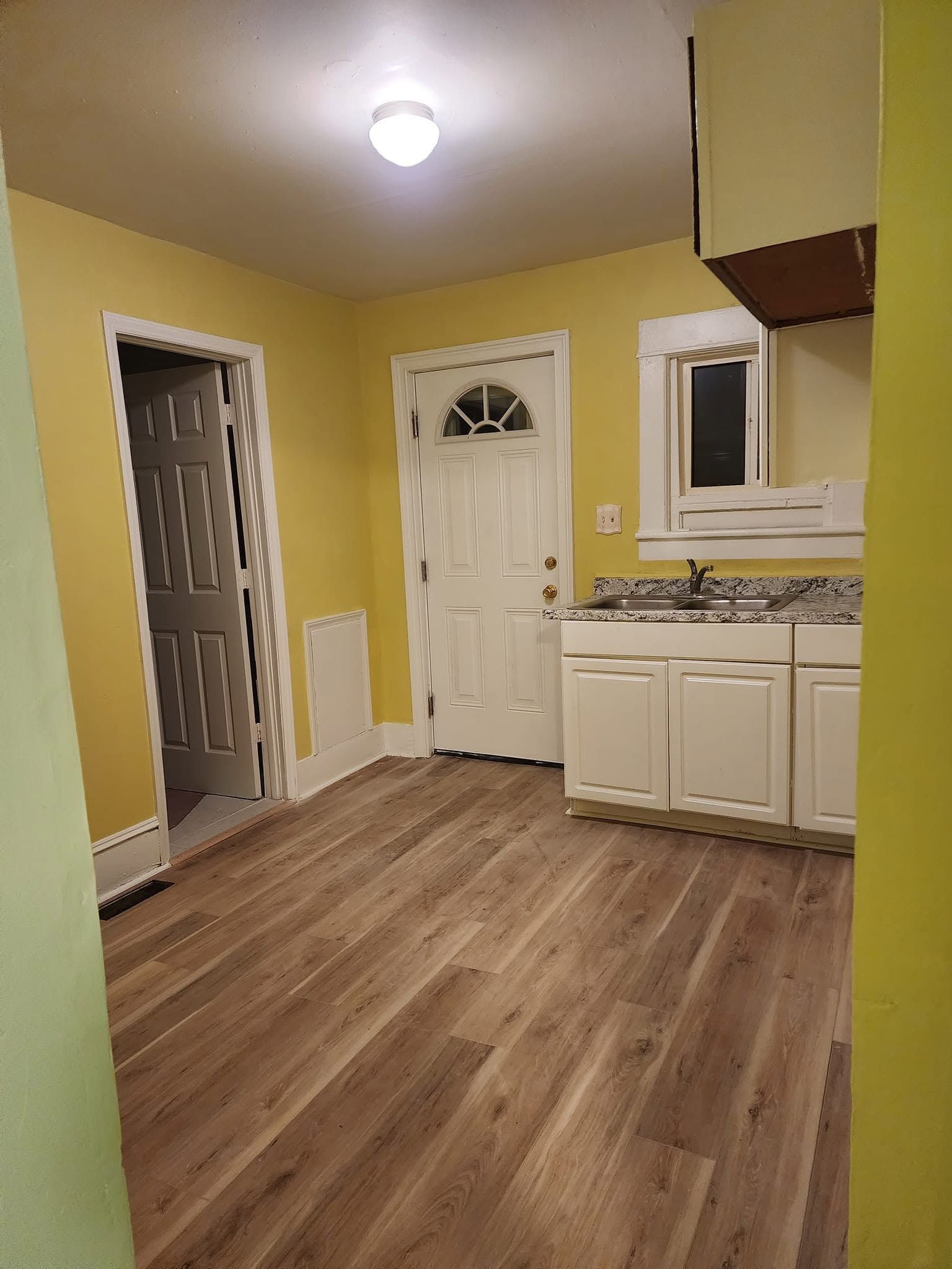 Empty yellow kitchen with wood-look floor, white cabinets, granite counter, door, and window.