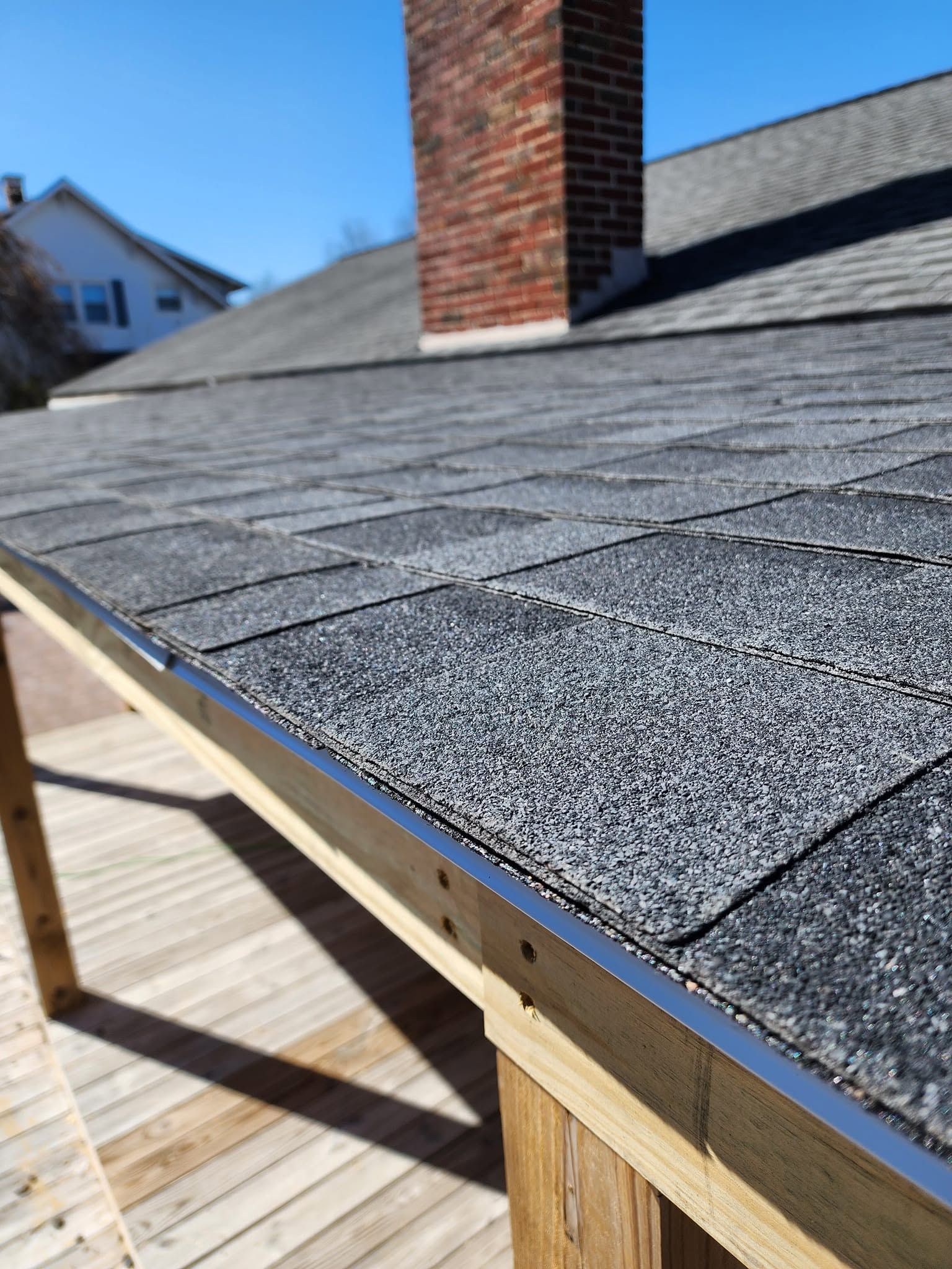 Close-up of a shingled roof edge with exposed trim and a brick chimney under blue sky