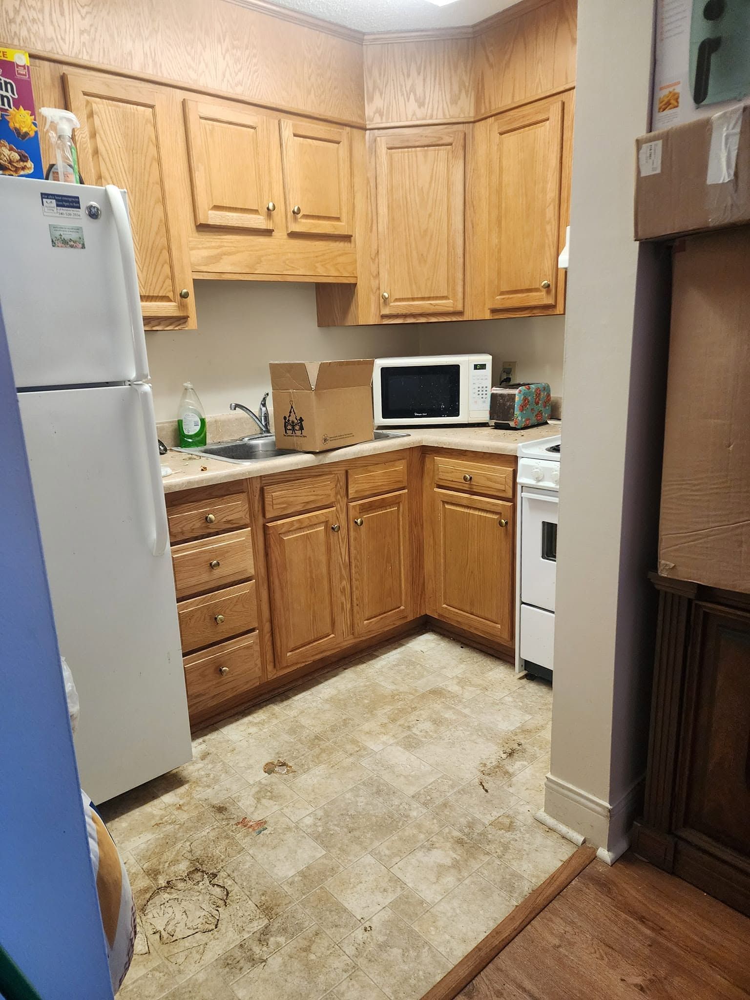 Small kitchen with light wood cabinets, white refrigerator, microwave, and worn beige linoleum floor.