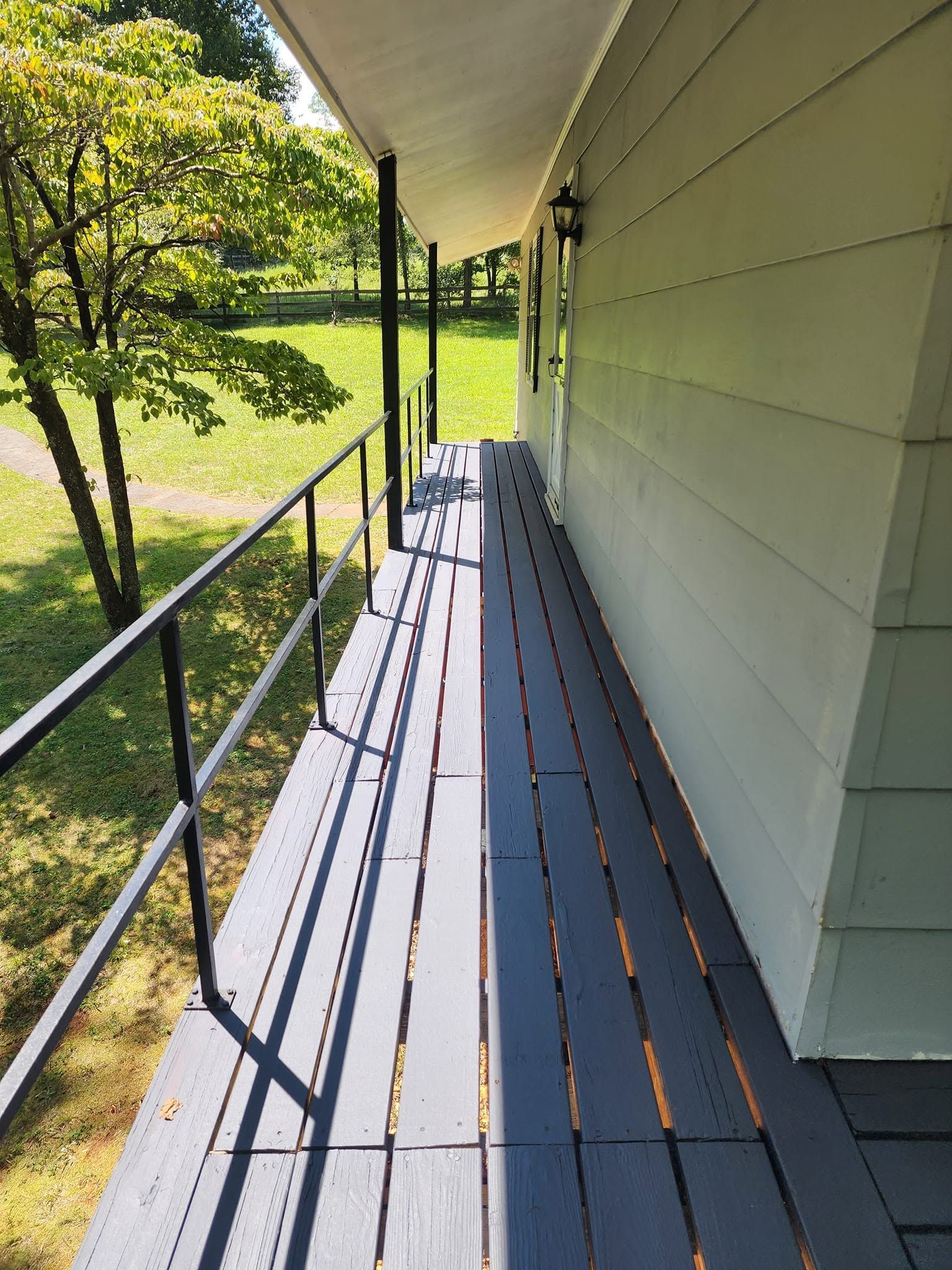 Covered apartment walkway with metal railing and sunlit wooden floorboards beside a grassy yard