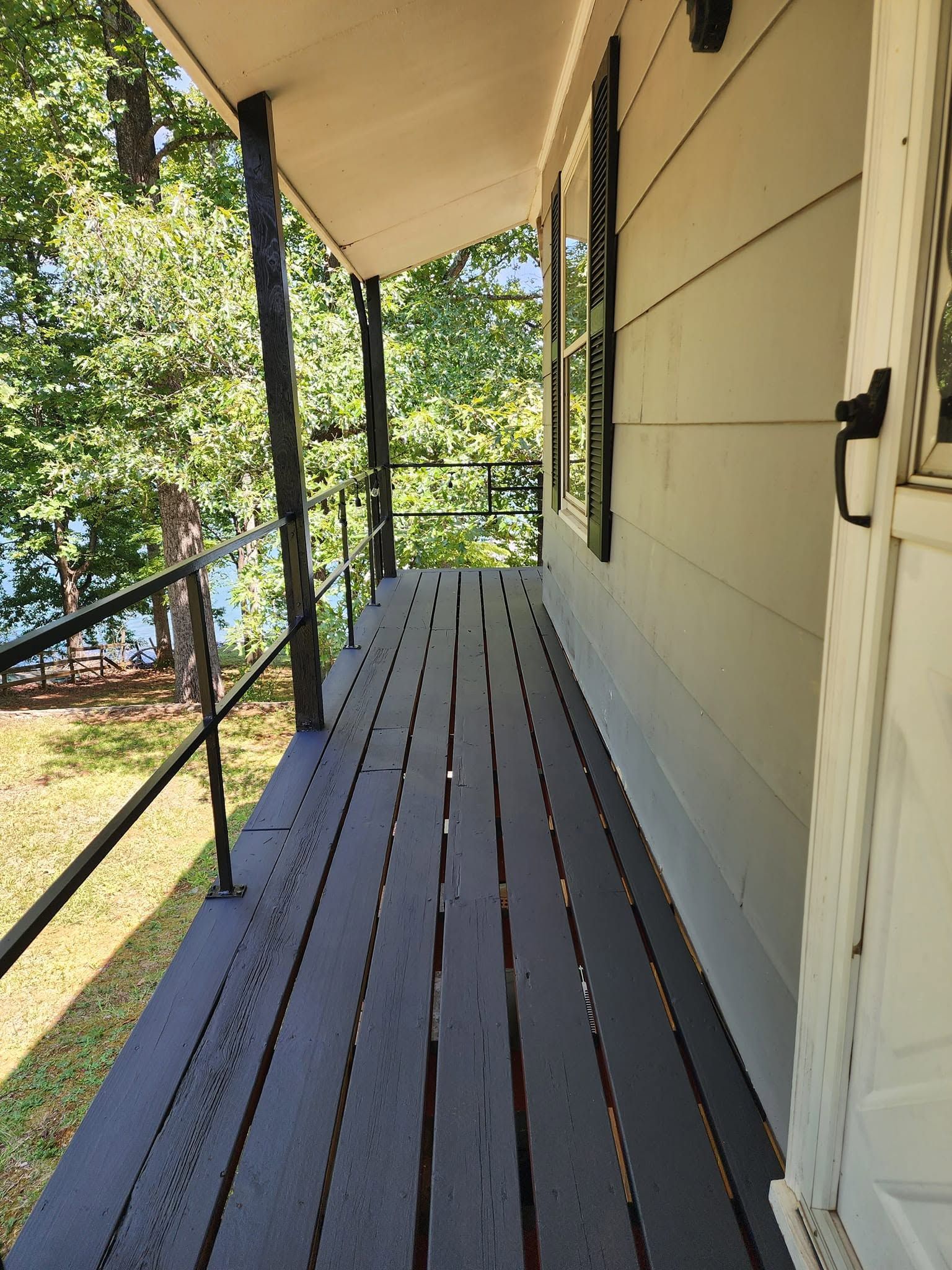 Covered wooden porch with black railing, beige siding, and trees overlooking a lake.