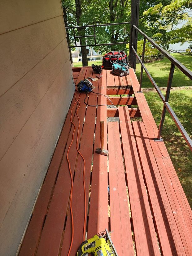 Red wooden porch with scattered tools and a yellow chainsaw, overlooking green trees and scaffolding.