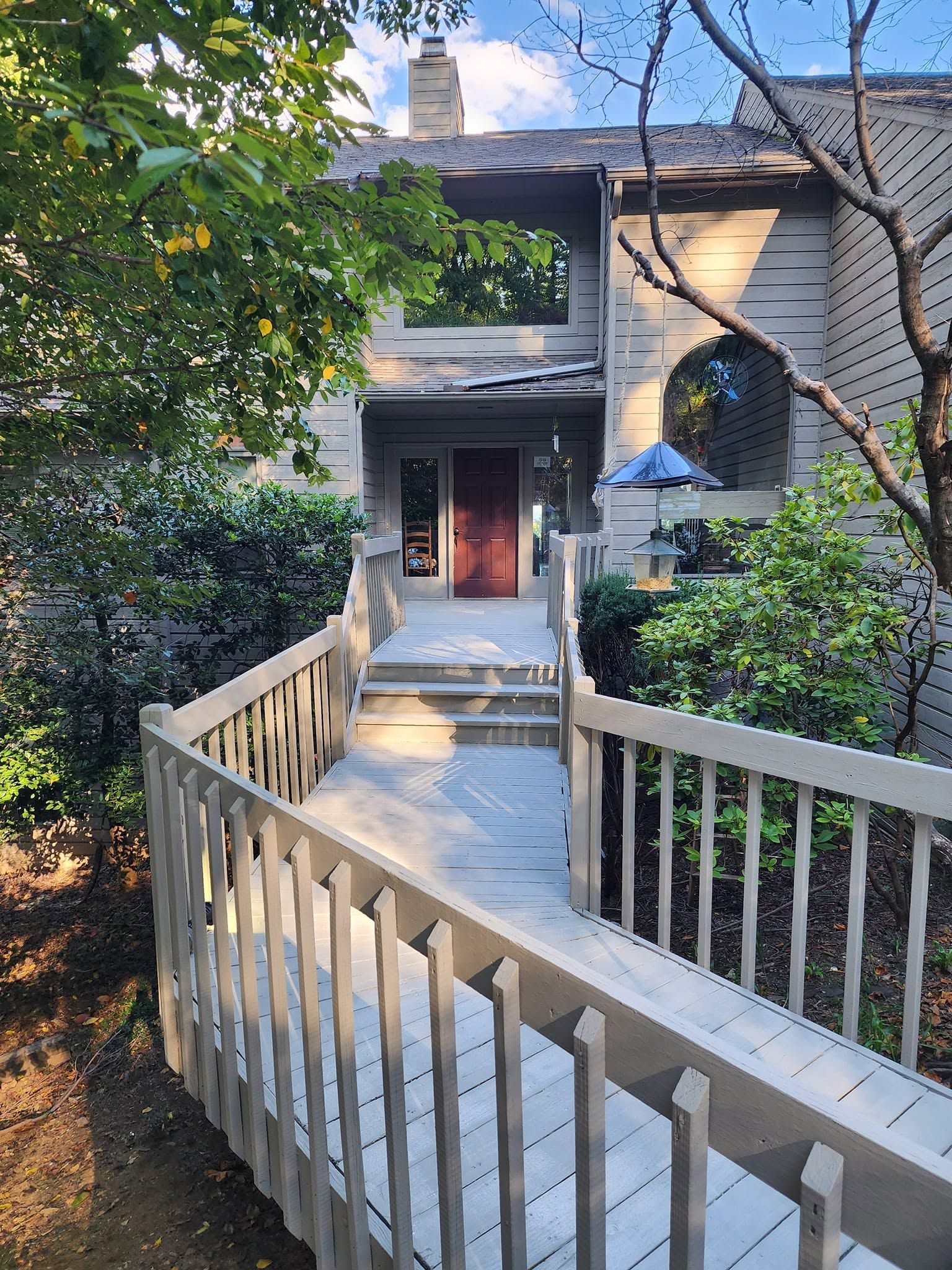 Curved wooden ramp leading to a house entrance with railings, greenery, and shaded porch.