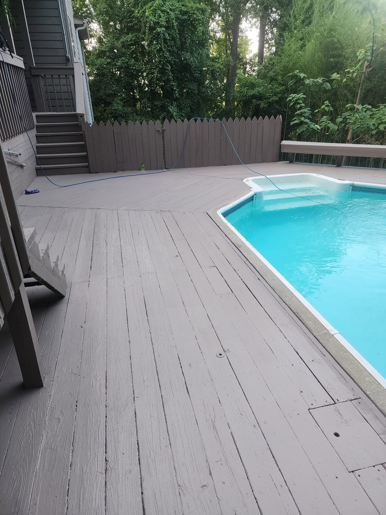 Wooden pool deck beside a turquoise swimming pool, with stairs and a fence lined by trees.