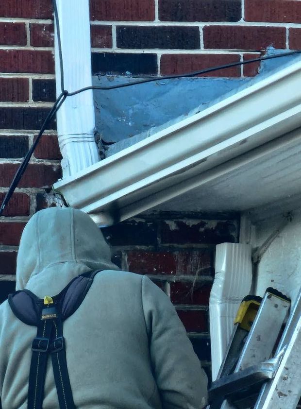 Person in gray hoodie inspecting a white gutter and downspout against a brick house wall.