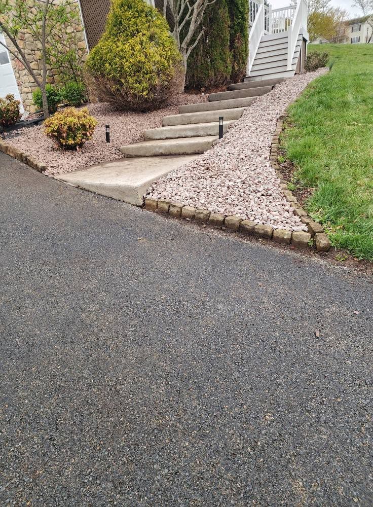 Front walkway with concrete steps, shrubs, and a gravel border beside a paved driveway.