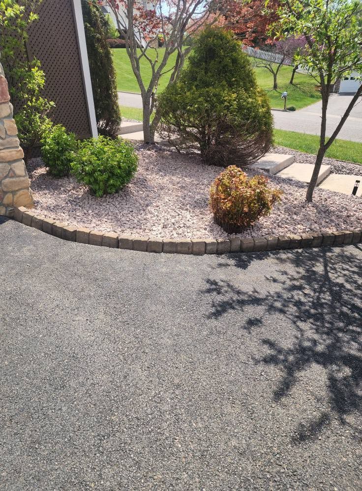 Gravel landscaping bed with shrubs and a tree beside a paved driveway and sidewalk