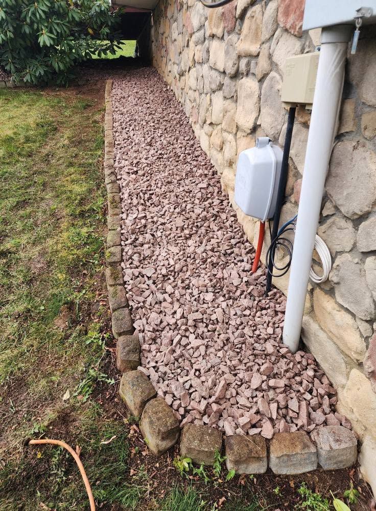 Rock-lined gravel drainage bed beside a stone house wall and utility pipes.