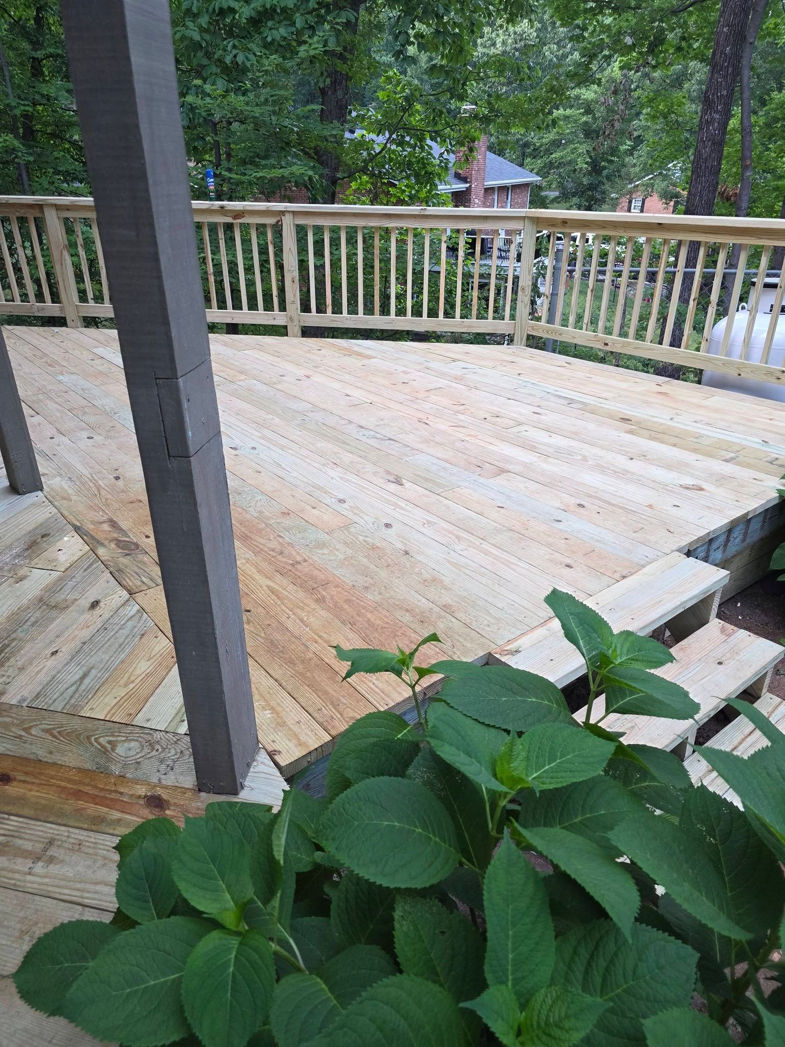Wooden deck with white railing and a dark post, viewed from a porch with green plants in the foreground.