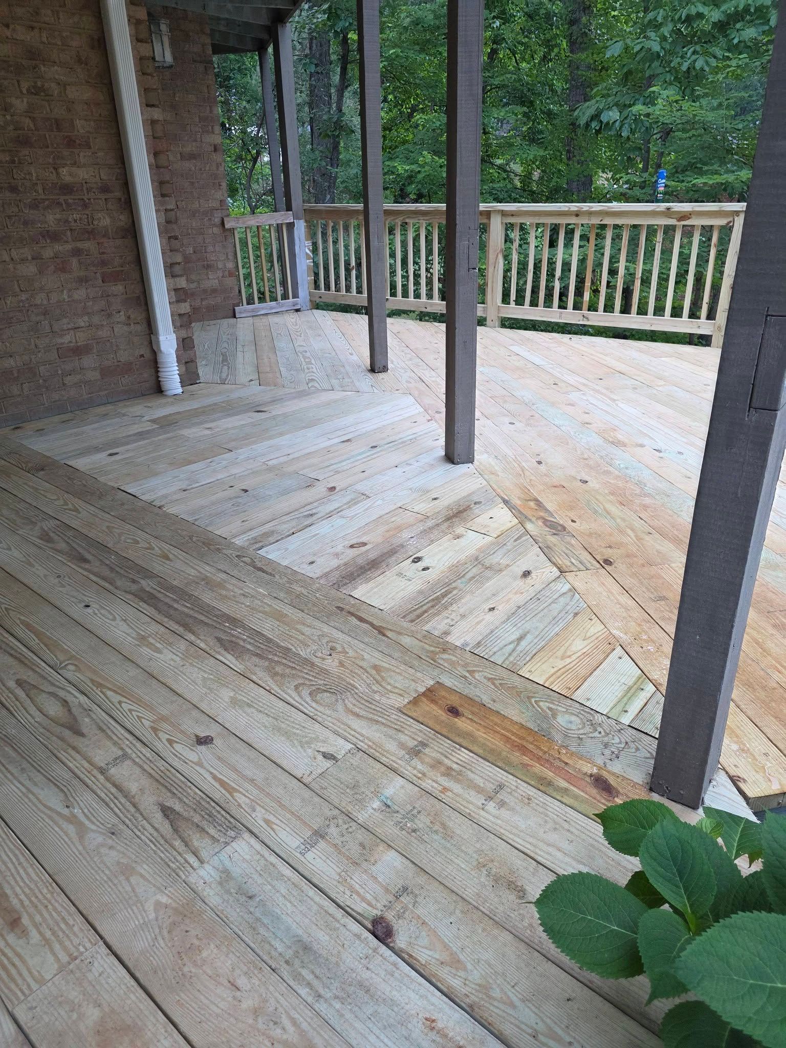 Wooden porch deck with railing, support posts, and a herringbone-pattern floor near a brick wall.