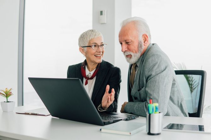 A man and a woman are sitting at a desk looking at a laptop.