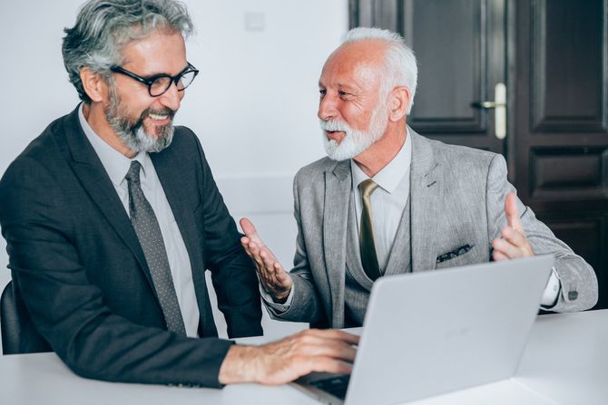 Two men are sitting at a table looking at a laptop computer.