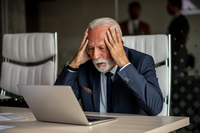 An older man is sitting at a desk looking at a laptop computer.
