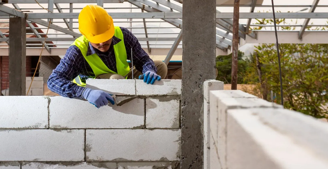 Construction worker laying concrete blocks on a building wall, wearing a yellow hard hat and gloves.