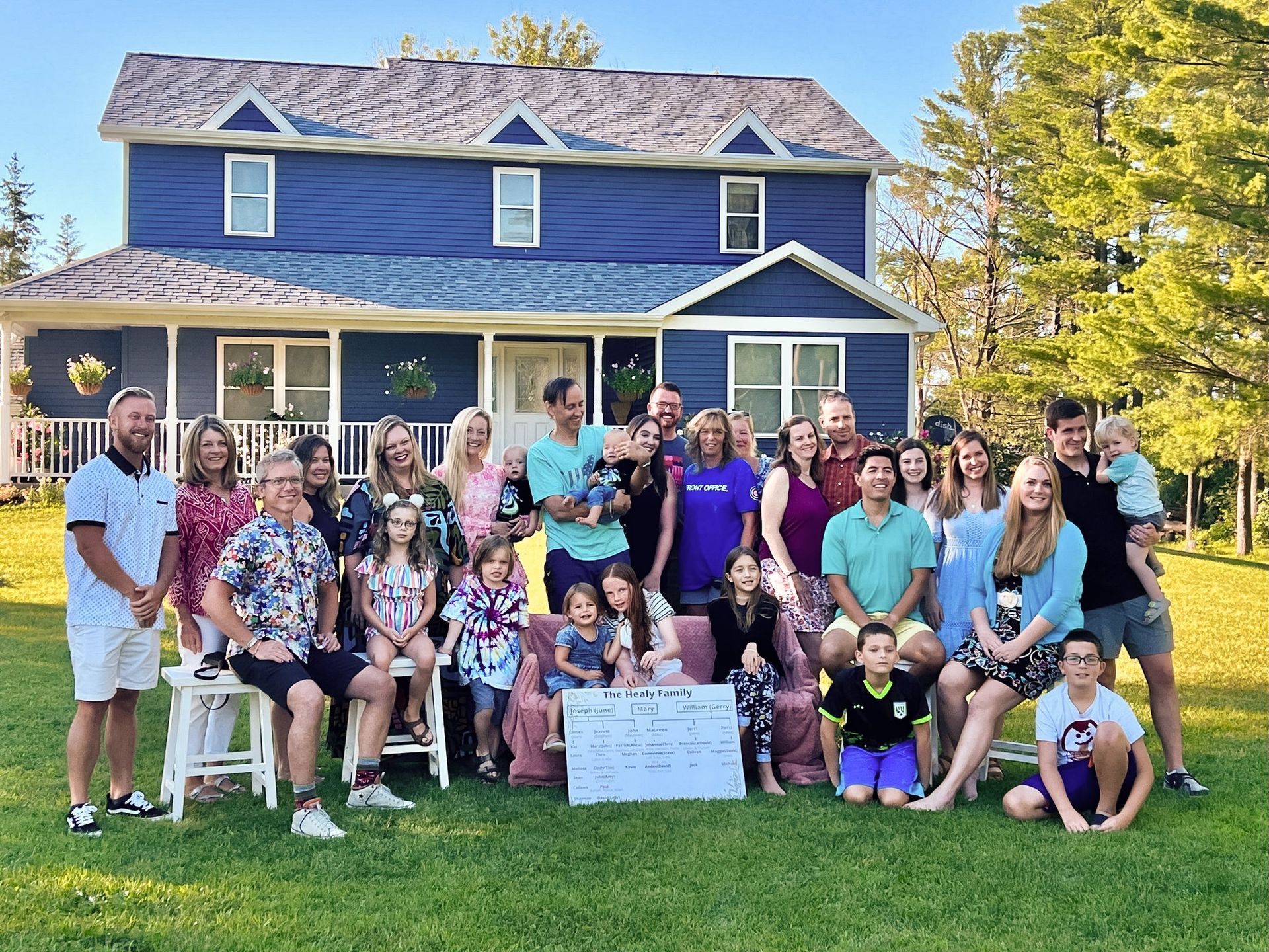 Large family group posing in front of a blue house, holding a sign in a grassy yard.