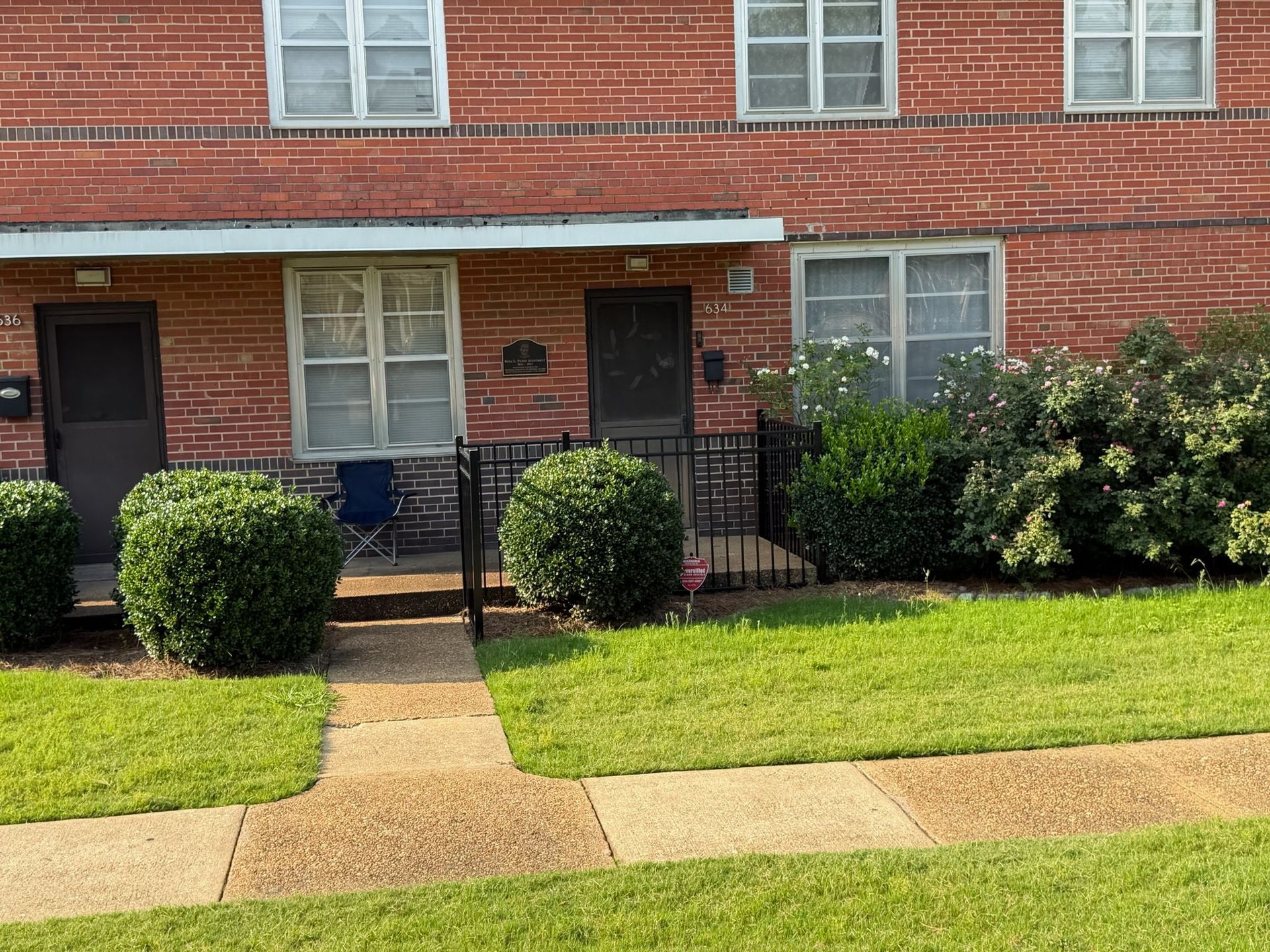 Brick apartment building with black doors, trimmed bushes, and a walkway in front of the lawn.
