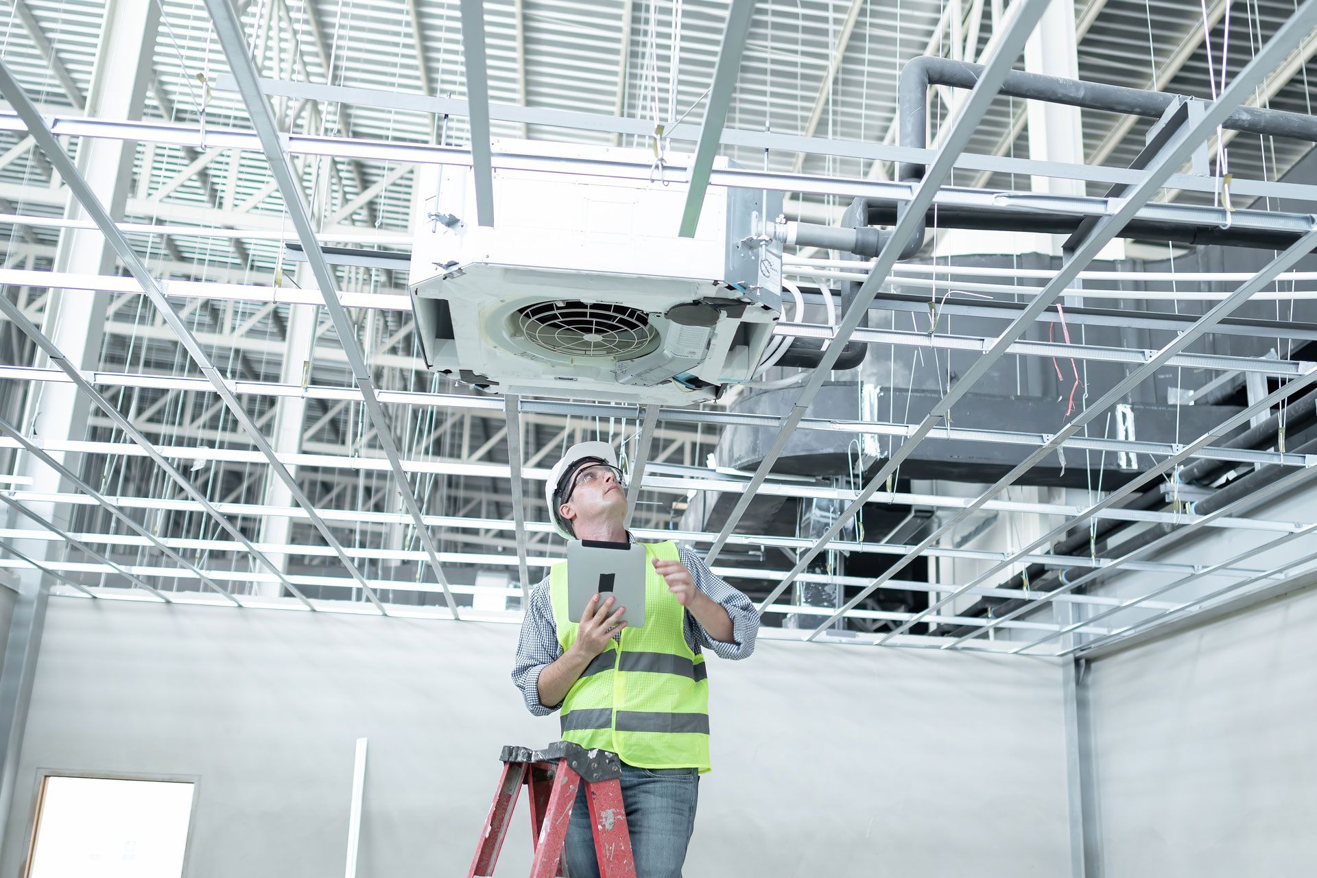 A construction worker is looking up at the ceiling of a building while using a tablet.